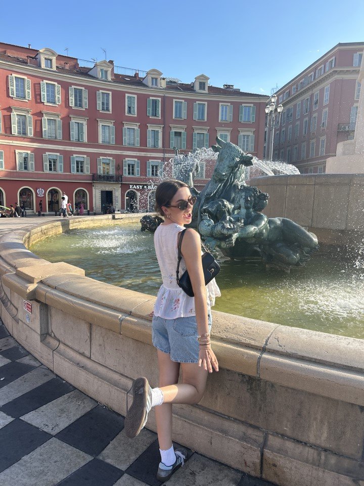A young woman with dark hair, wearing sunglasses, a white sleeveless top, denim shorts, white socks, and sneakers, posing with one leg bent at the knee and her foot resting on a stone fountain's edge, in an urban square with a bronze sculpture of horses and water sprays, and colorful buildings in the background on a bright, sunny day.
