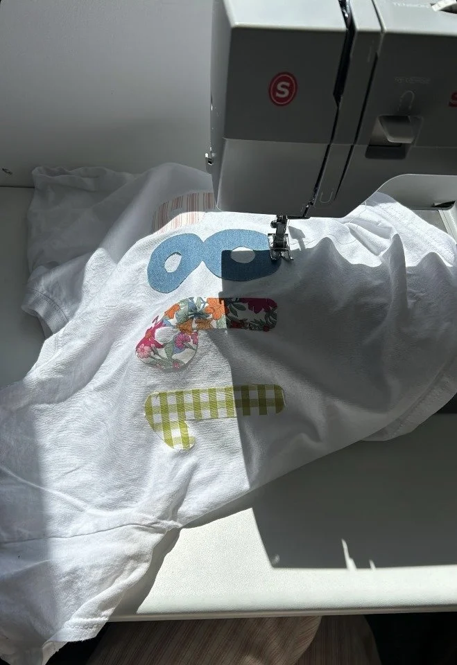A sewing machine stitching a colorful fabric appliqué design onto a white T-shirt with floral and checkered patterns.