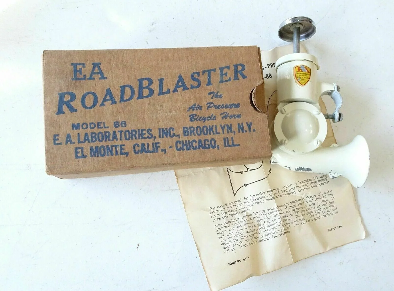 A vintage E. A. Roadblaster air pressure bicycle horn with a white body and a metal handle, displayed next to its original cardboard box and an instruction sheet.