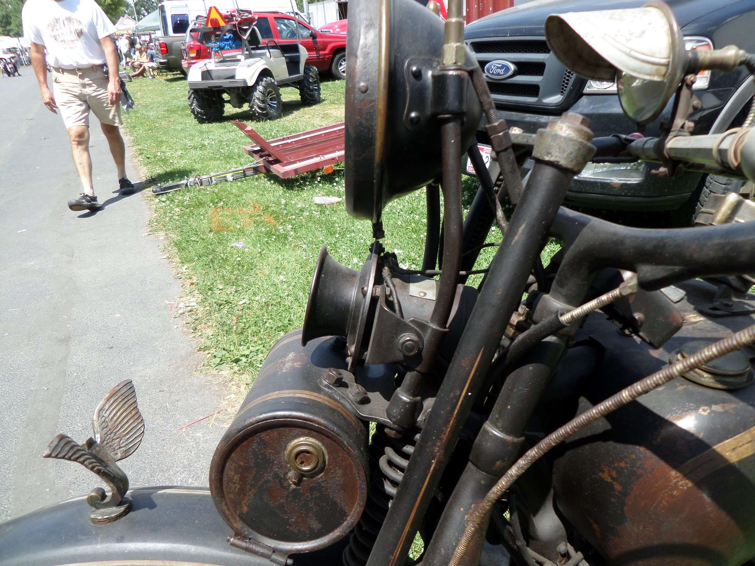Close-up of an old, rusty motorcycle's front section, showing the headlight, handlebar, and part of the front wheel and fender. In the background, a man walks past cars and a small red utility vehicle with a trailer attached at an outdoor event.