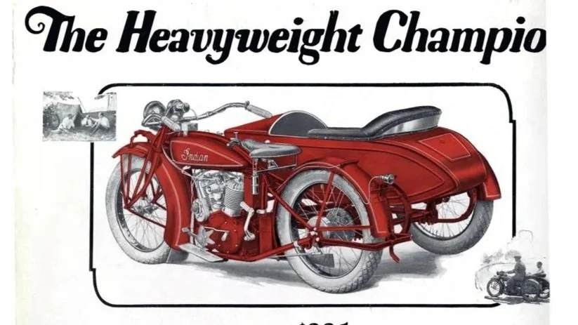 Vintage red motorcycle with a sidecar labeled 'Indian' against a white background, with the title 'The Heavyweight Champion' and small photos of motorcycle racing and riding.