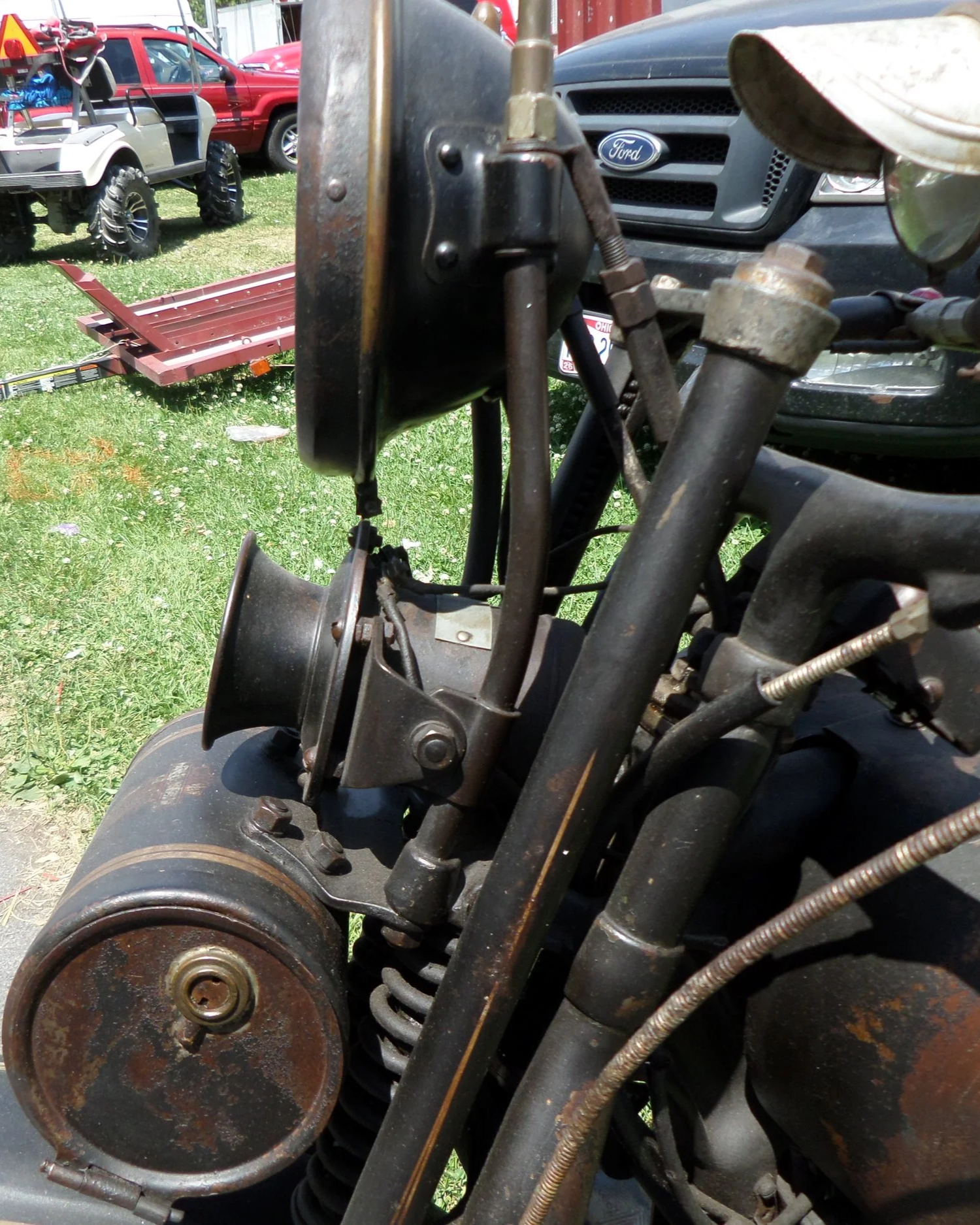Close-up of an old, rusty motorcycle front end, including the headlight, suspension, and wiring, with vehicles and grass in the background.