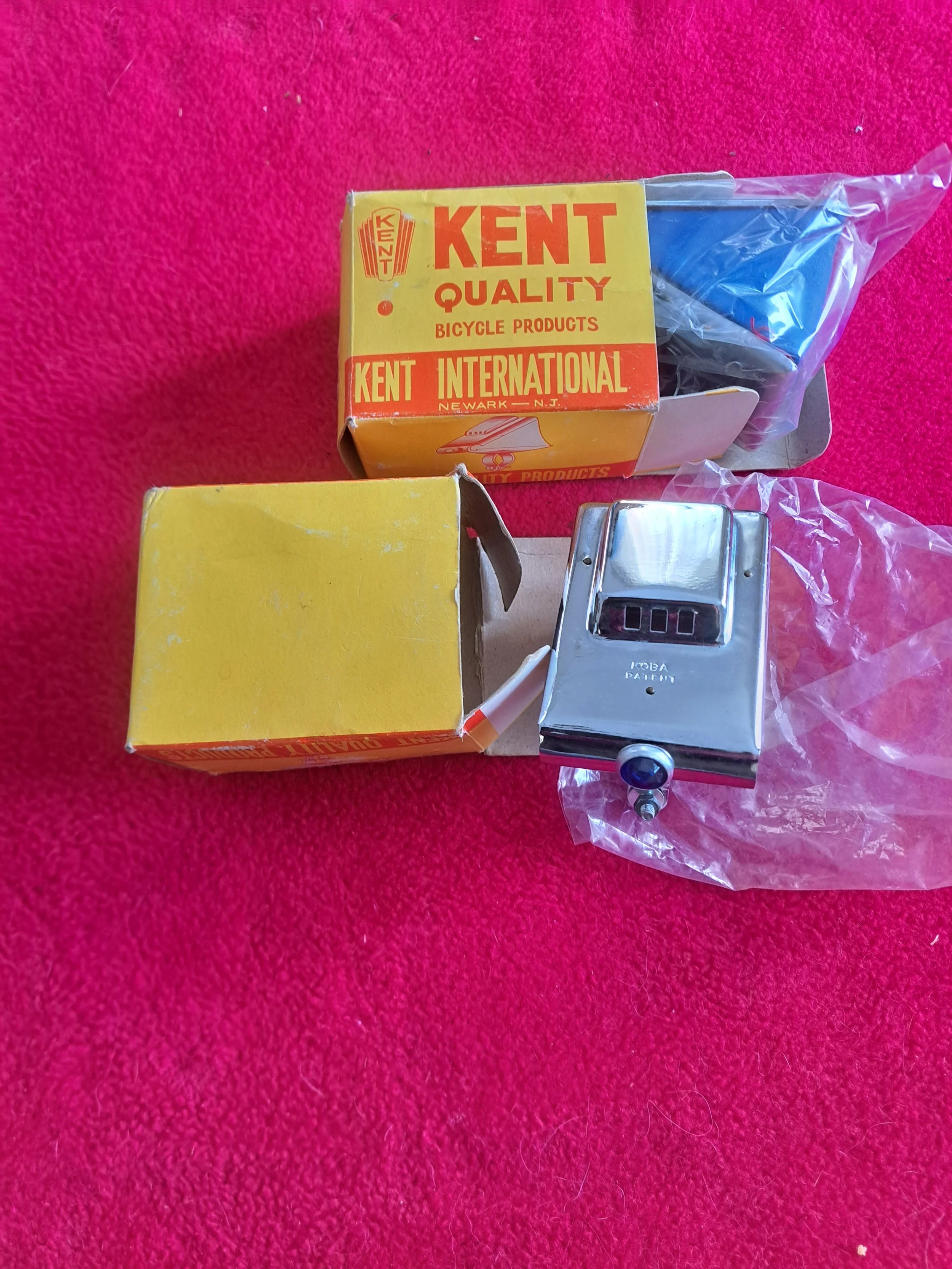 A metal bicycle hub with a small black knob and a gauge, along with two yellow boxes, one open with plastic bag, on a red textured surface.