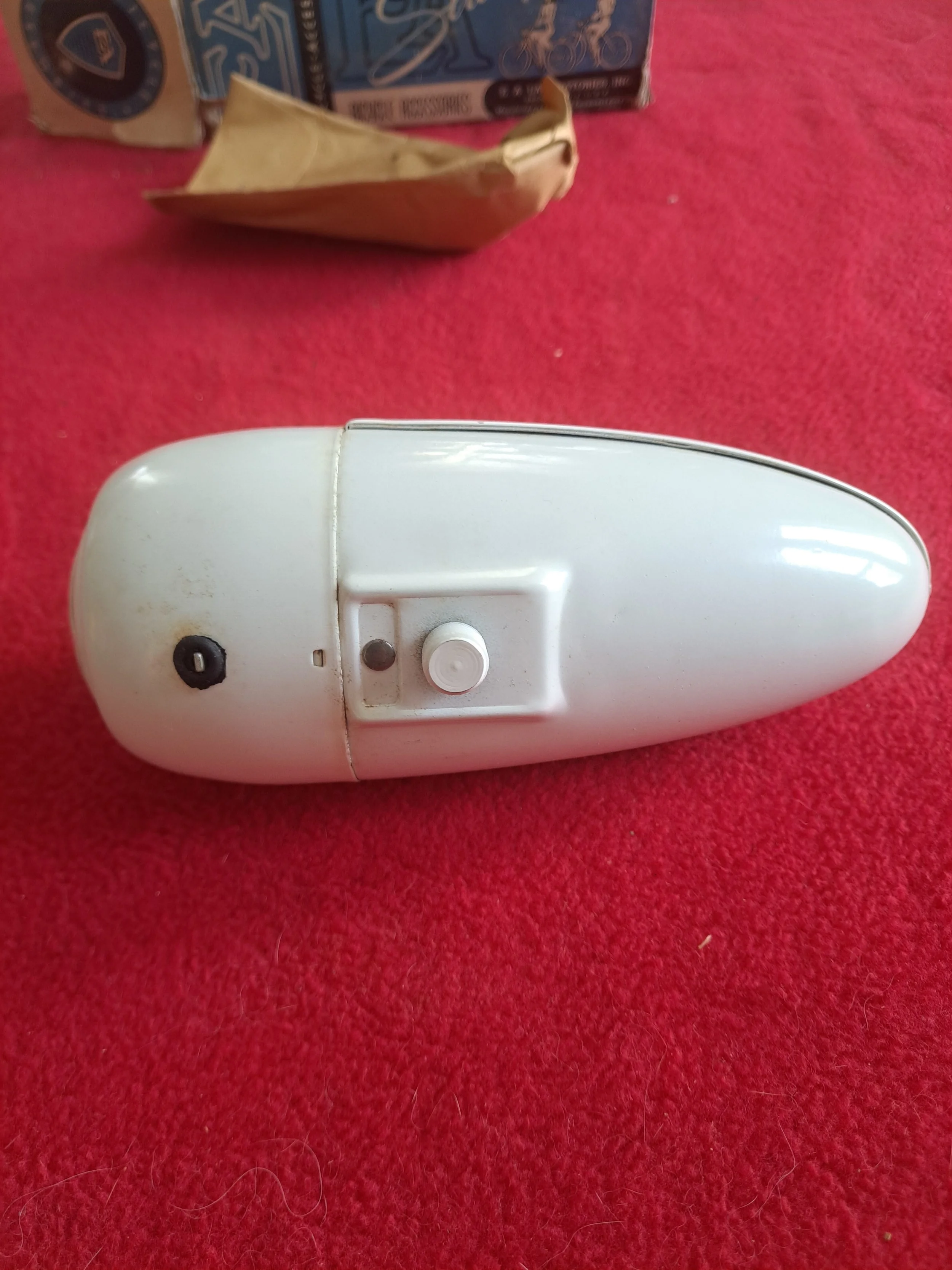 A vintage white electric shaver lying on a red textured surface, with a brown paper bag and a boxed toy in the background.