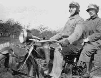 Two men in vintage clothing sitting on a motorcycle or moped outdoors