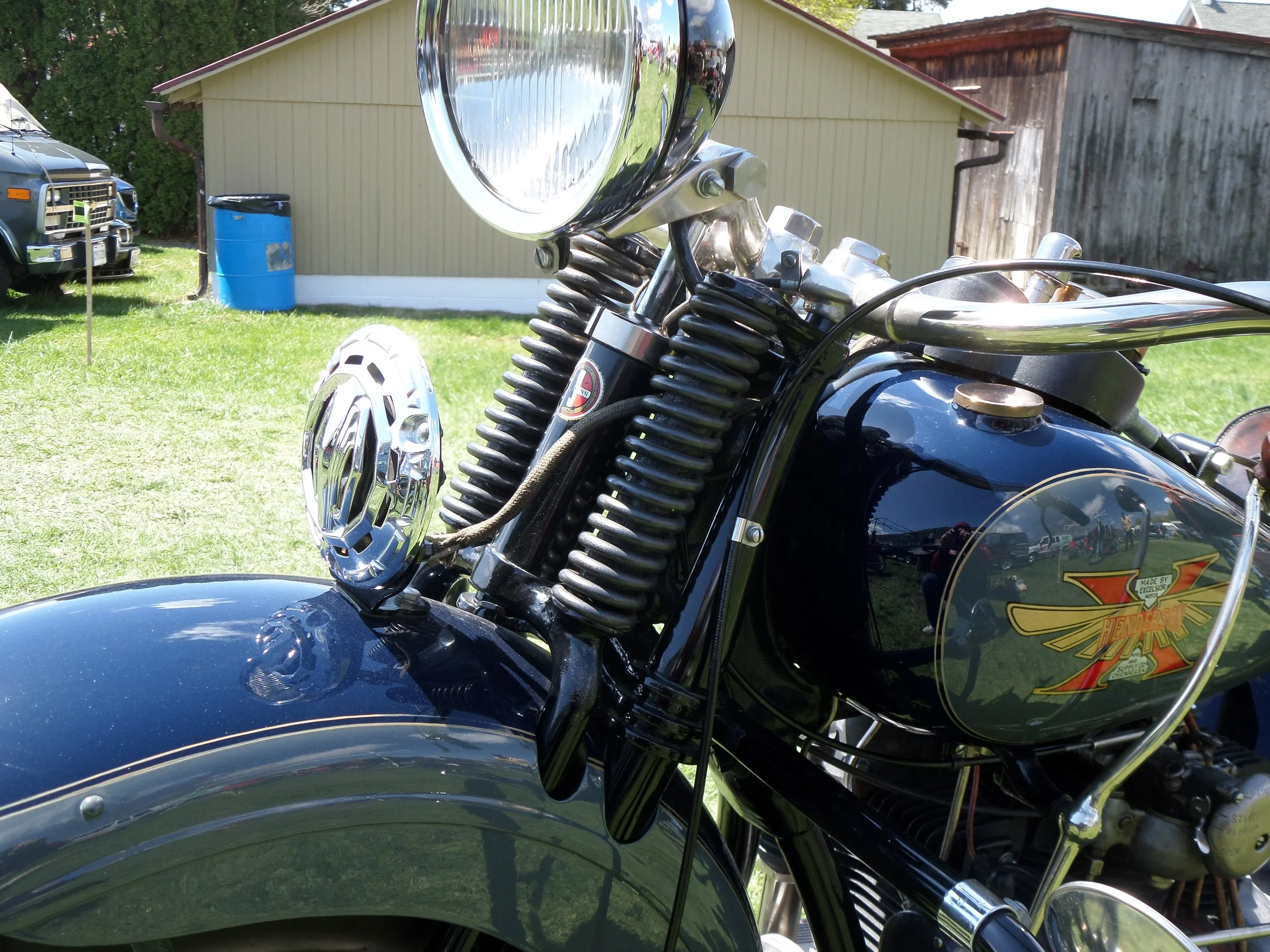 Close-up of a vintage black motorcycle with a large round headlight, springs on the front fork, and a bright yellow and red 'Henderson' logo on the gas tank, with a grassy yard and a shed in the background.