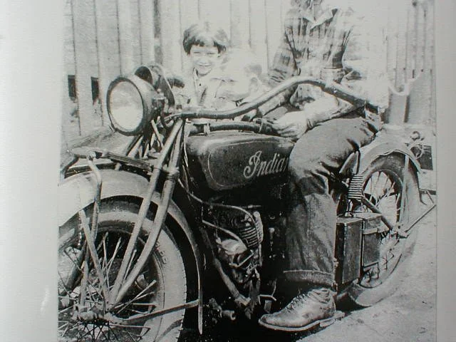 Black and white photo of two boys sitting on a vintage Indian motorcycle, with a wooden fence in the background.