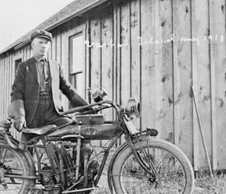 A man in vintage clothing riding an old-fashioned motorcycle in front of a wooden building.