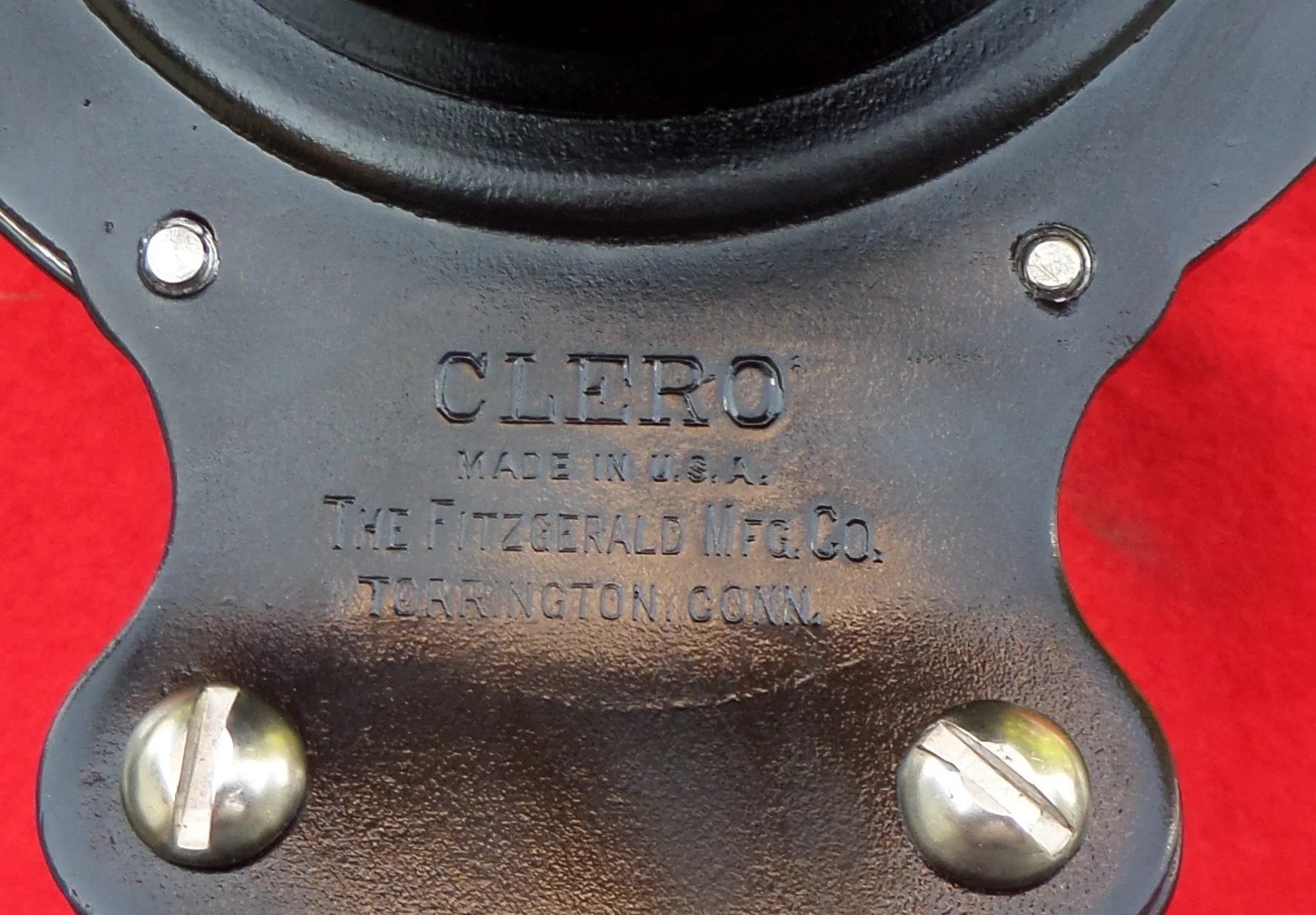 Close-up of a black metal object with the brand 'CLERO' and the text 'Made in U.S.A. The Fitzerald Mfg. Co. Tarrantin G.N.' engraved on it, with silver screws attaching it to a red background.