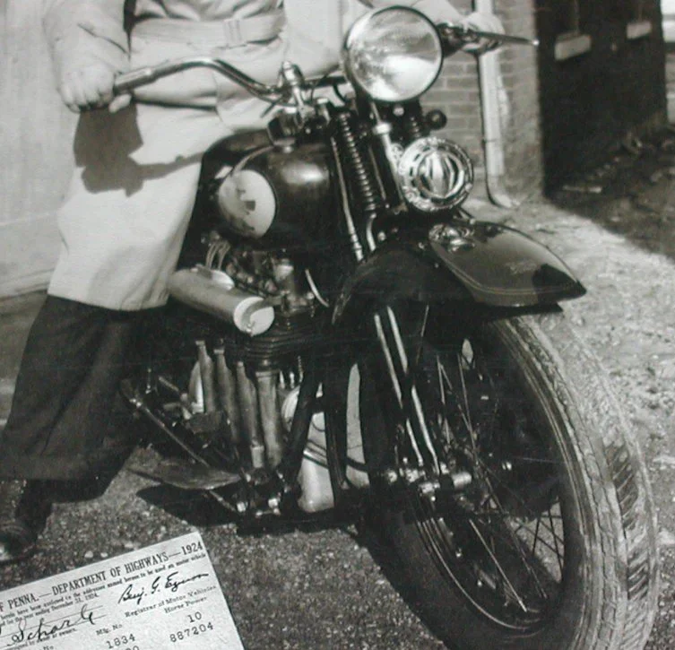 Black and white photo of a person standing next to a vintage motorcycle, with a document in the foreground. The motorcycle has a large front headlight and a prominent fuel tank.