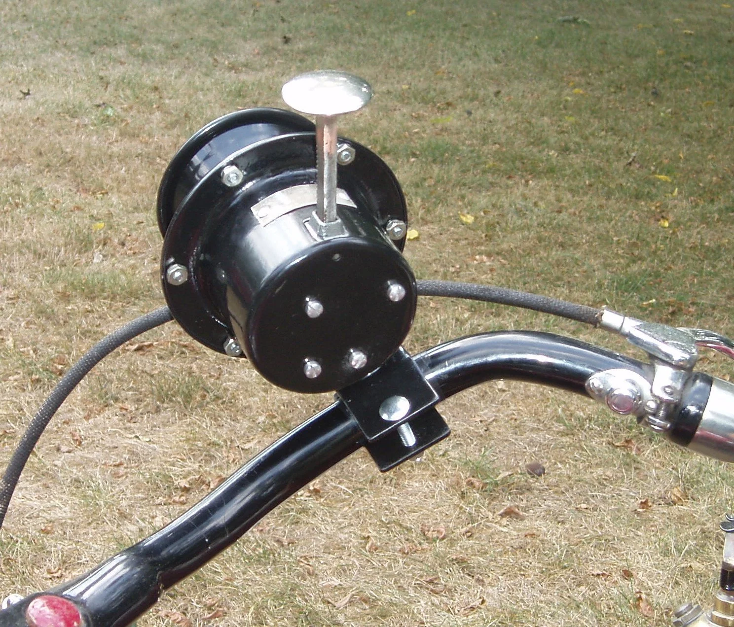A close-up of a black motor or generator mounted on the handlebar of a bicycle or motorcycle, with visible bolts, wires, and a metallic handle on a grassy outdoor surface.