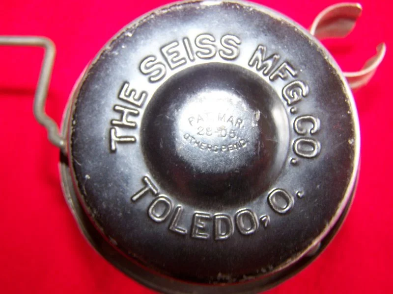 Close-up of a black metal object with embossed writing, including 'THE SEISS MFG. CO. TOLEDO, O.' and a central circular indentation with smaller embossed text 'PAT. MAR. 28-05, OTHERS PEND' on a red background.