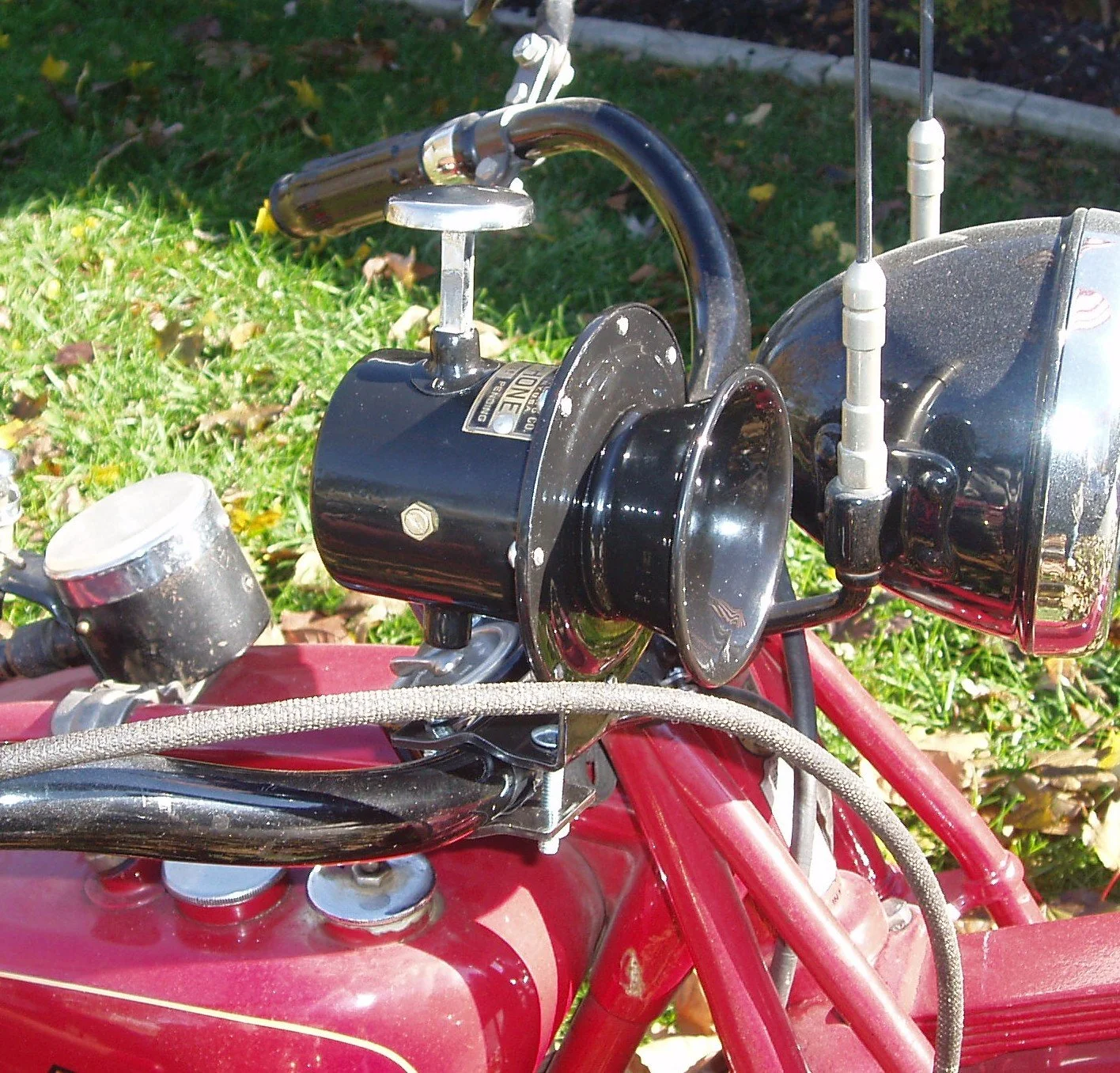 Close-up of a vintage motorcycle handlebar and headlight on a red frame, with a grassy background.