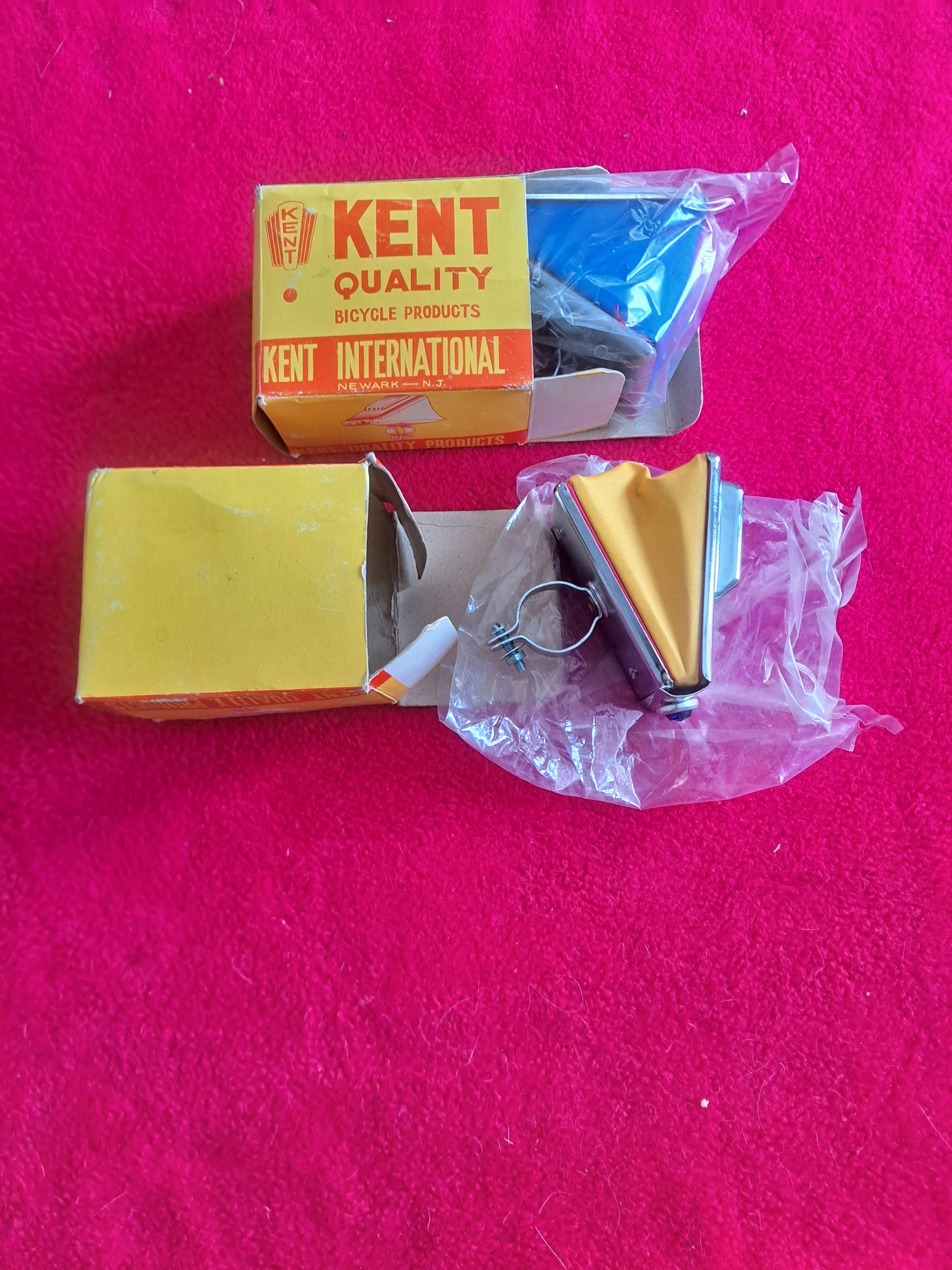 Vintage bicycle road reflector with yellow reflective surface and attached metal wire, along with two vintage bike parts boxes on a pink surface.