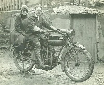 Two people riding a vintage motorcycle together, both wearing coats and hats, in front of a construction site or dirt area.