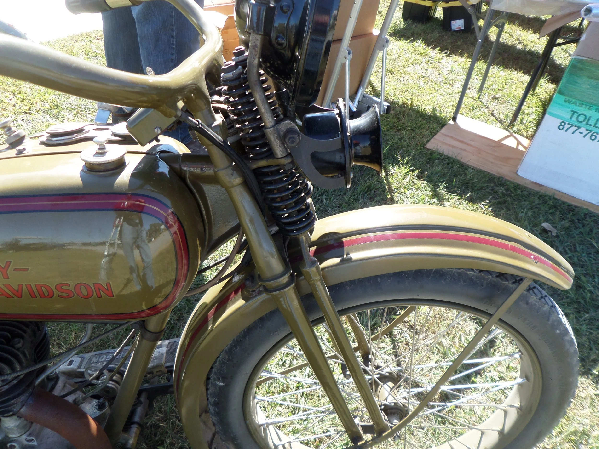 Close-up of a vintage gold and black Harley-Davidson motorcycle, focusing on the front wheel, fender, and part of the gas tank, with a street and parking lot in the background.