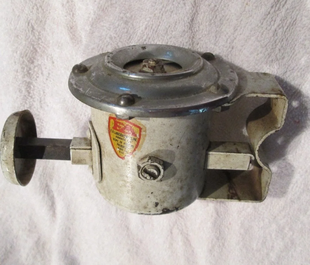 An old metal meat grinder with a handle on the side, a rotating metal plate on top, and a large opening on the side for inserting meat, placed on a light-colored fabric background.