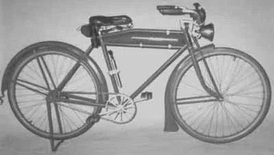 Black and white photo of an old-fashioned bicycle with a large front wheel and smaller rear wheel, featuring a storage box on top and a shield mounted on the front.