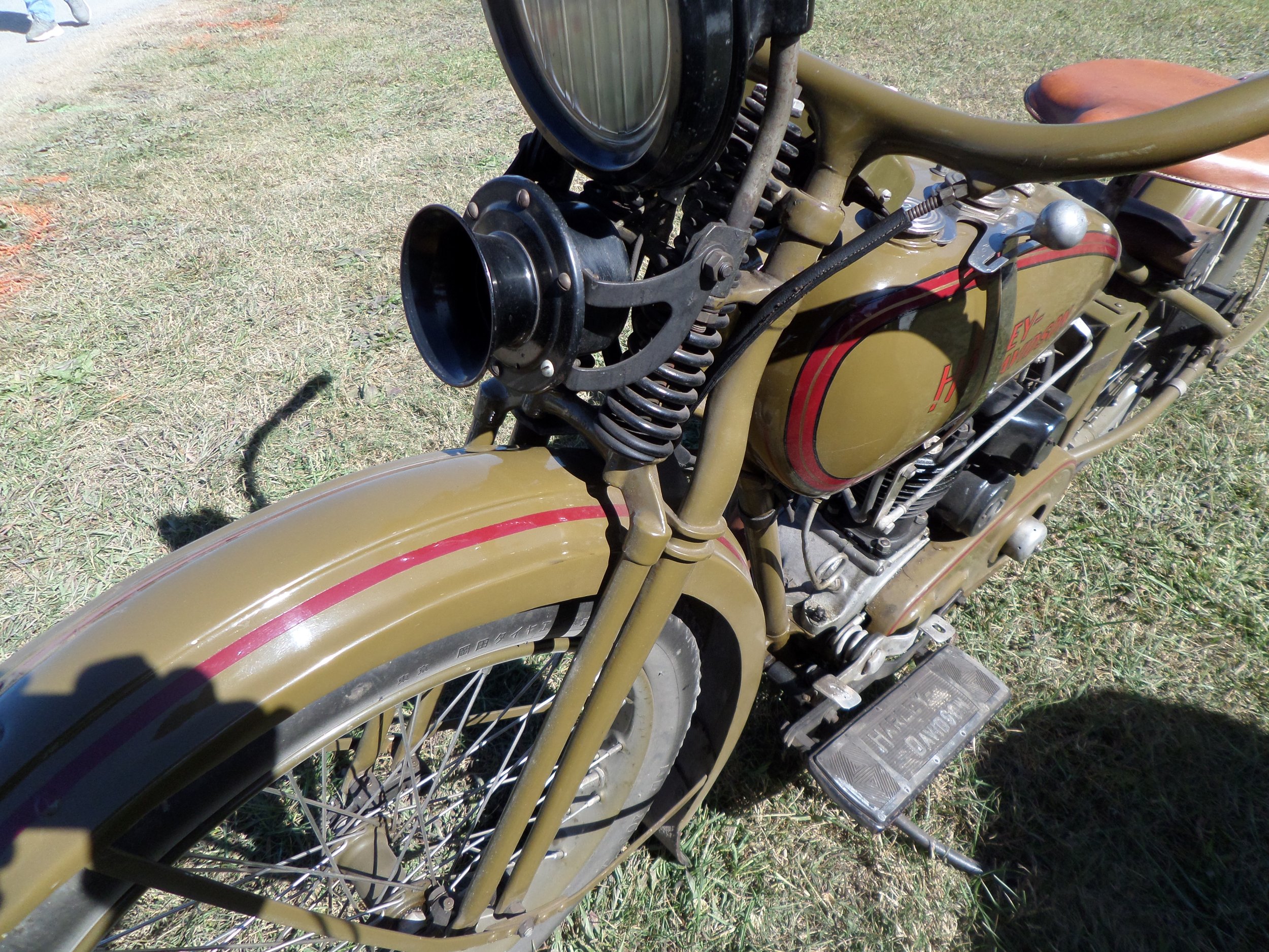 Close-up of a vintage military-style motorcycle with a khaki green body and red pinstripes, parked on grass.