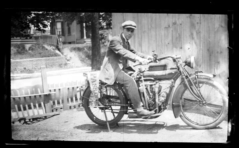 A young man in vintage clothing and a cap sitting on a classic motorcycle in an outdoor setting.