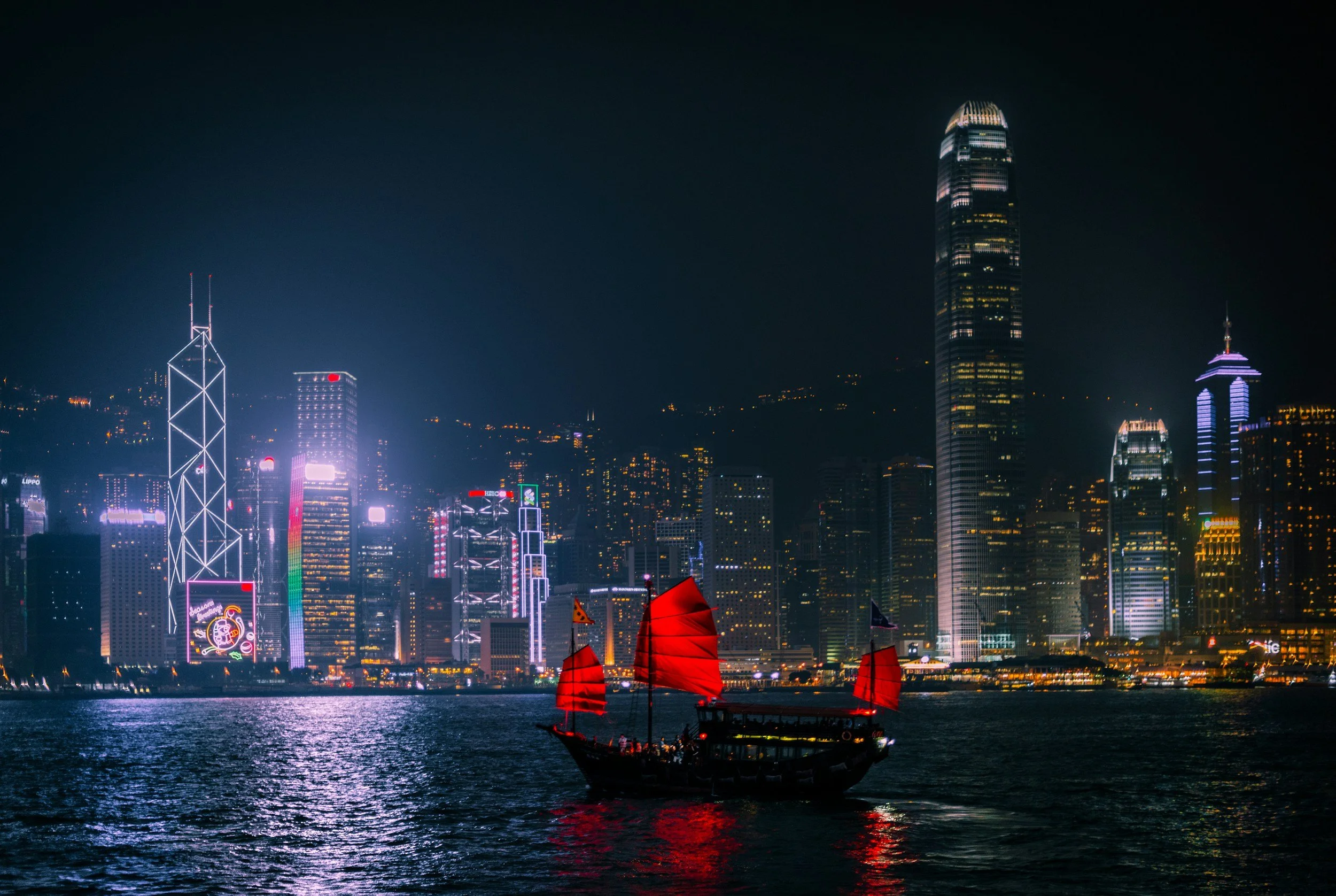 A traditional Chinese junk boat with red sails sailing on the water at night, with a brightly lit city skyline featuring skyscrapers and high-rise buildings in the background.