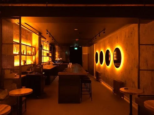 Dimly lit modern bar with circular wall art and a long bar counter on the left. Warm ambient lighting creates an inviting atmosphere.