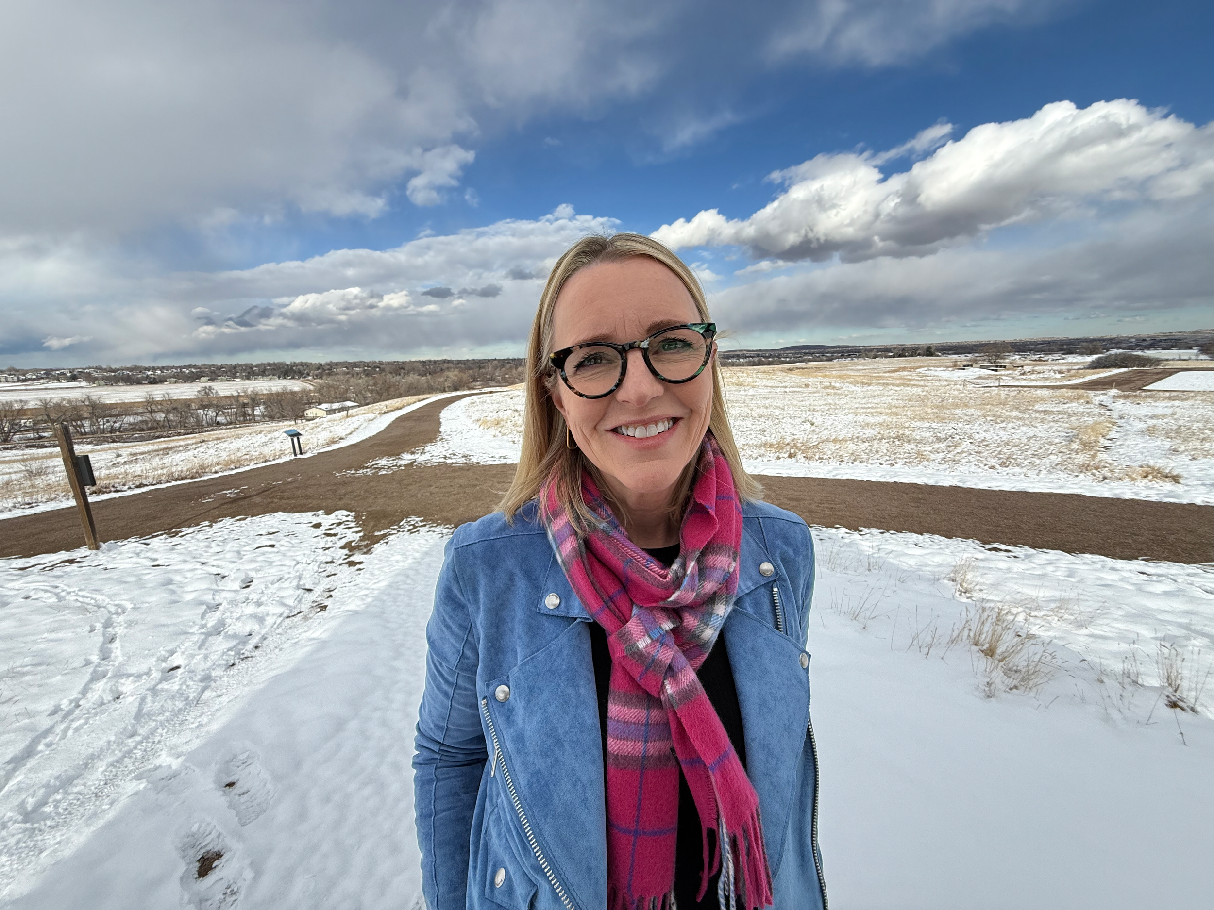 A woman with blonde hair, glasses, and a bright smile outdoors in a snowy landscape. She is wearing a blue jacket and a pink plaid scarf, with snow-covered fields and a cloudy sky in the background.