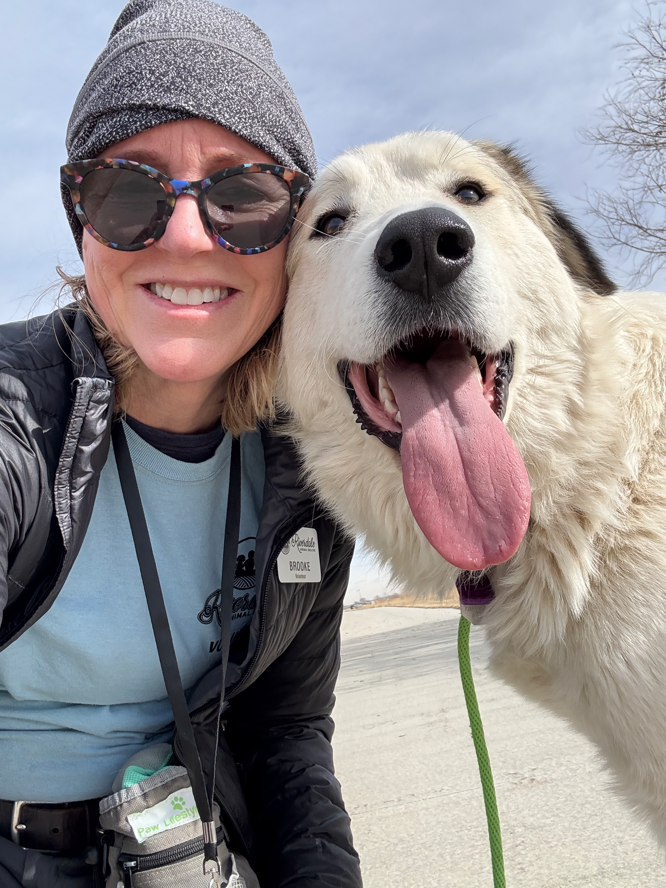 A woman and a large, fluffy white Great Pyrenees rescue dog taking a selfie outdoors with a cloudy sky and leafless trees in the background.