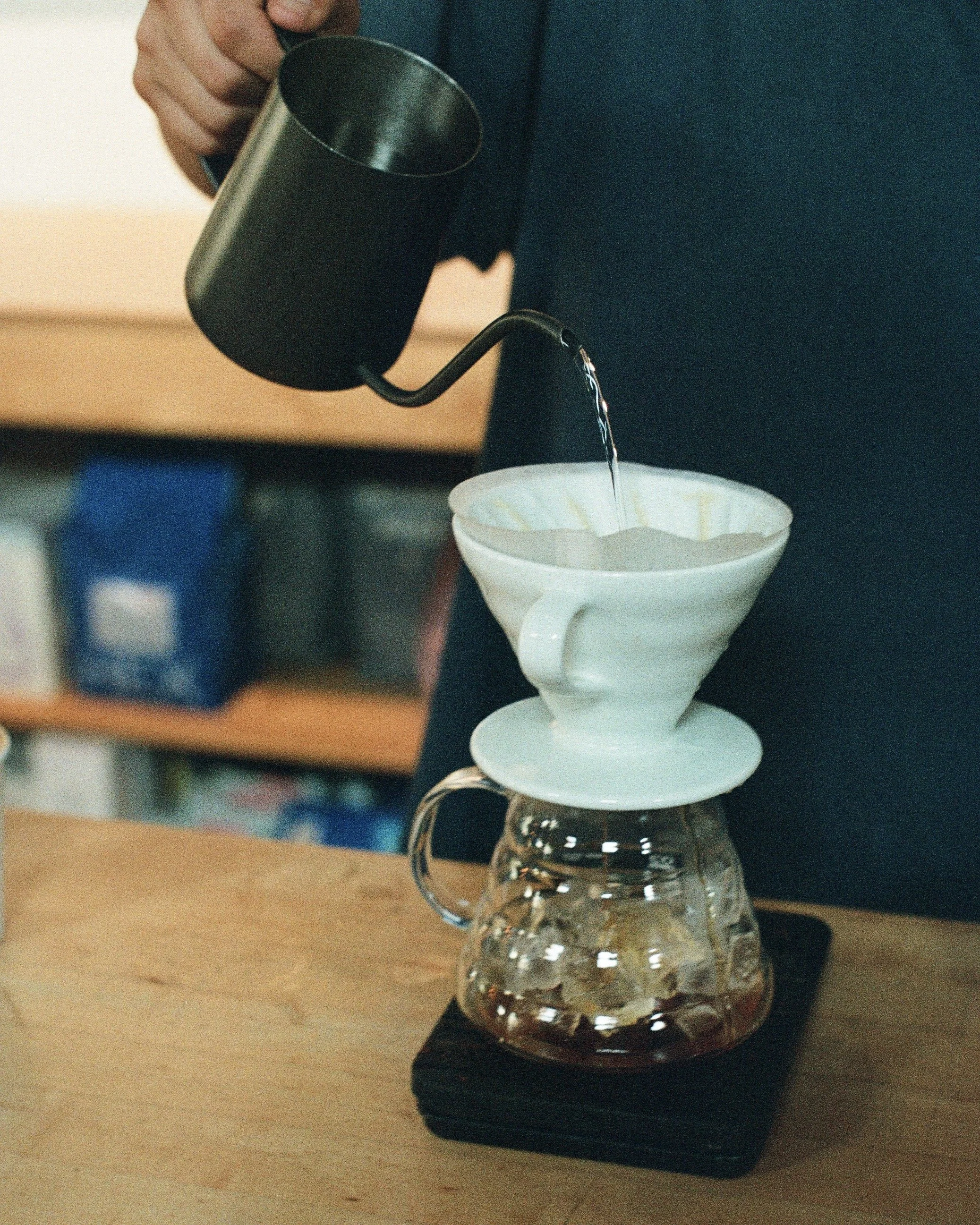 A person is pouring hot water from a black kettle through a white ceramic dripper filled with coffee grounds, which is placed on top of a glass carafe on a wooden surface.