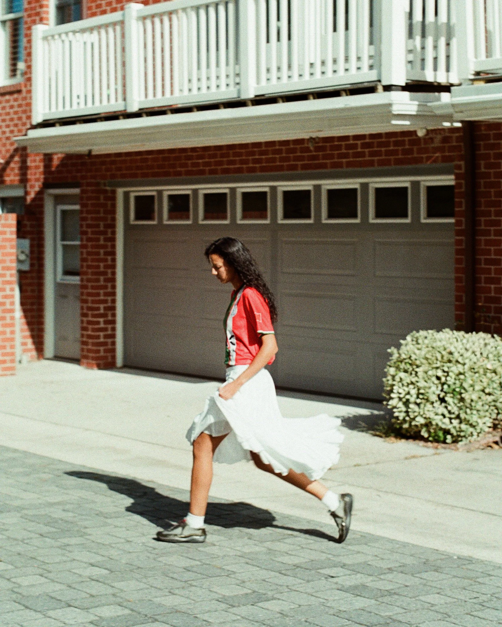 A woman with curly dark hair is walking on a sidewalk in front of a brick building with a garage door. She is wearing a red shirt, a white skirt, and sneakers, with a white jacket flowing behind her.