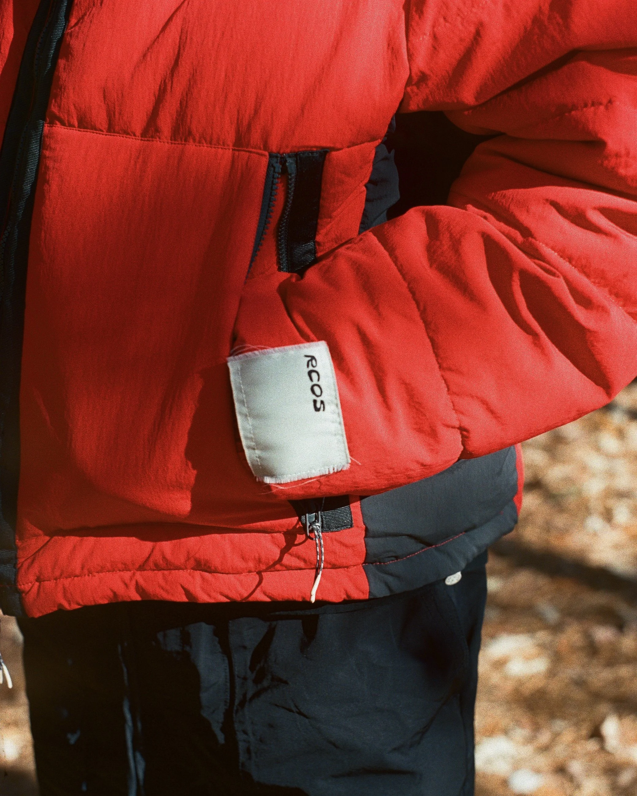 Close-up of a person wearing a red REI Co-op insulated jacket and black pants, standing outdoors with a blurred natural background.