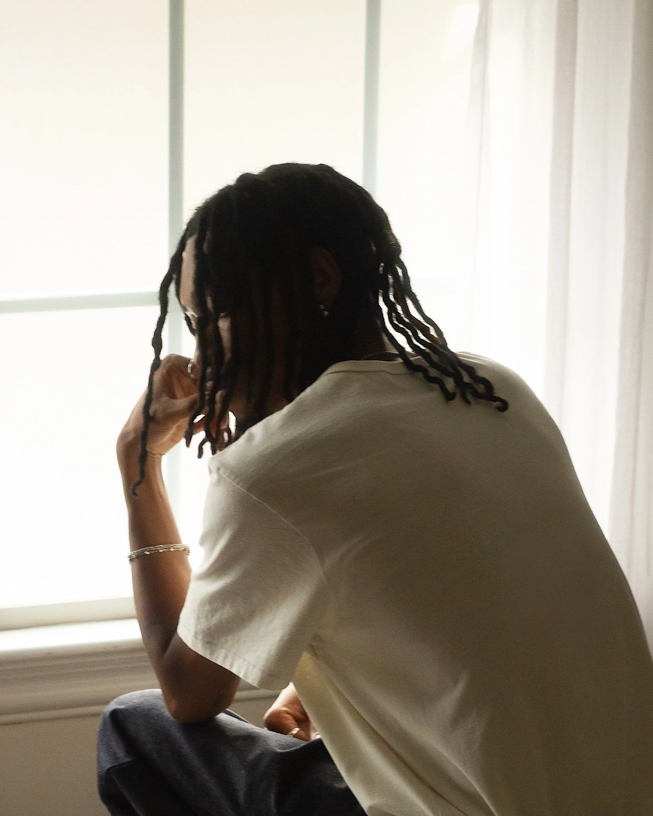 Person with long dreadlocks sitting by a window, resting their head on their hand.