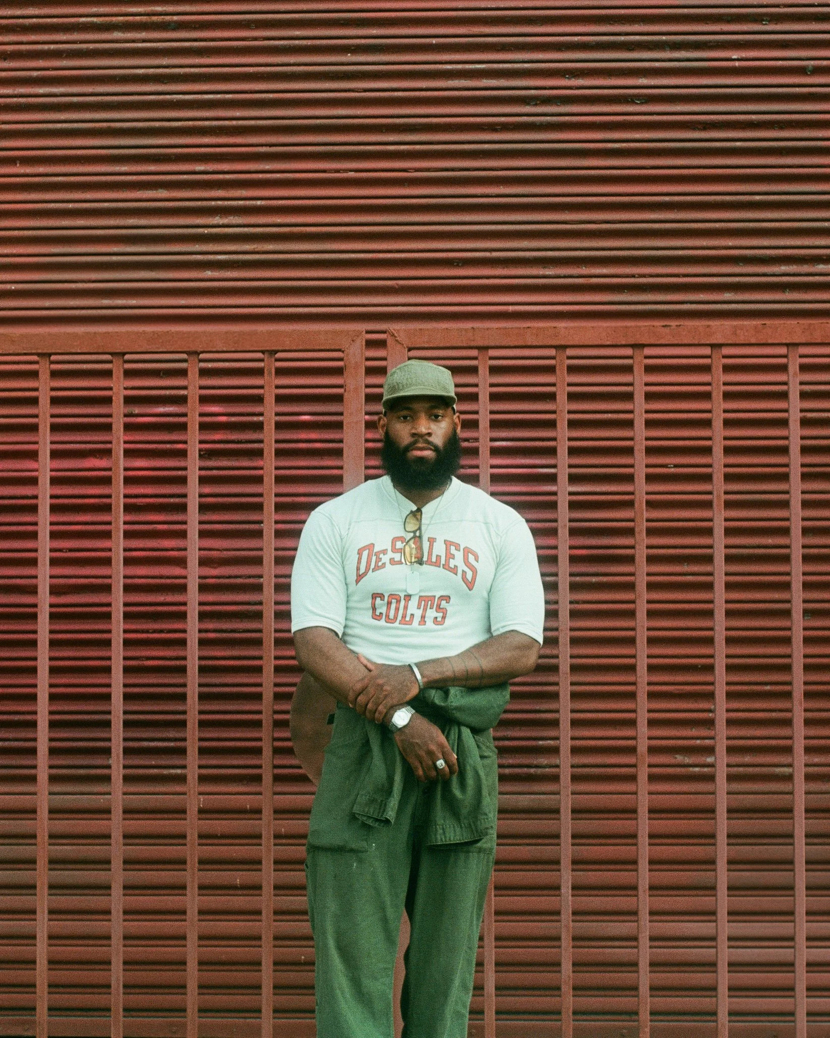 A man with a beard standing in front of a red metal fence wall, wearing a gray cap, sunglasses hanging on his shirt, a white T-shirt with 'De-SLIES Colts' written on it, green pants, and holding a green jacket.