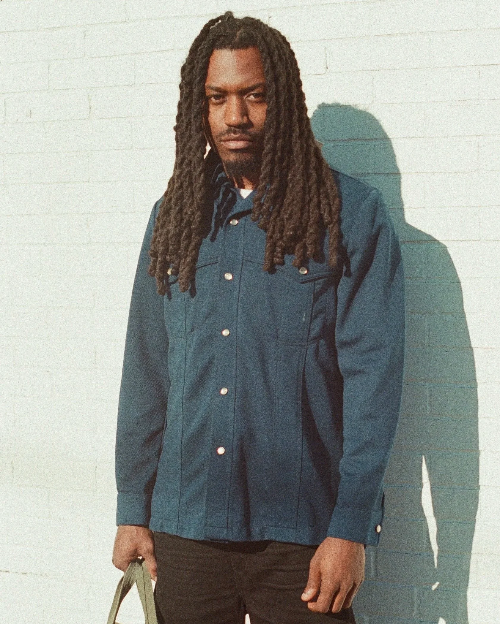 A young man with long dreadlocks wearing a dark denim jacket standing against a white brick wall with sunlight casting a shadow behind him.