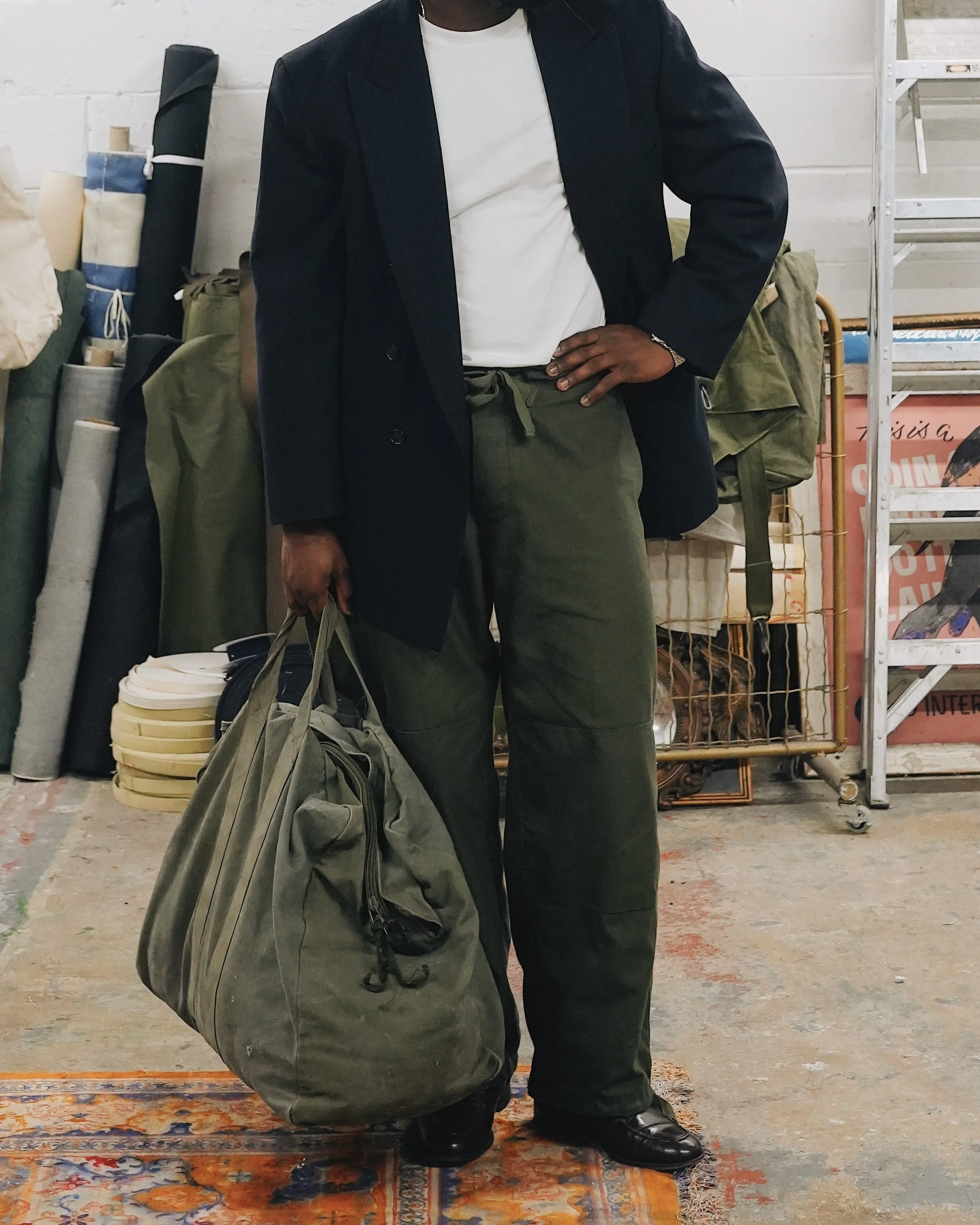 Person standing in front of a cluttered background, holding a large green duffel bag, wearing dark pants, a white t-shirt, and a black blazer.