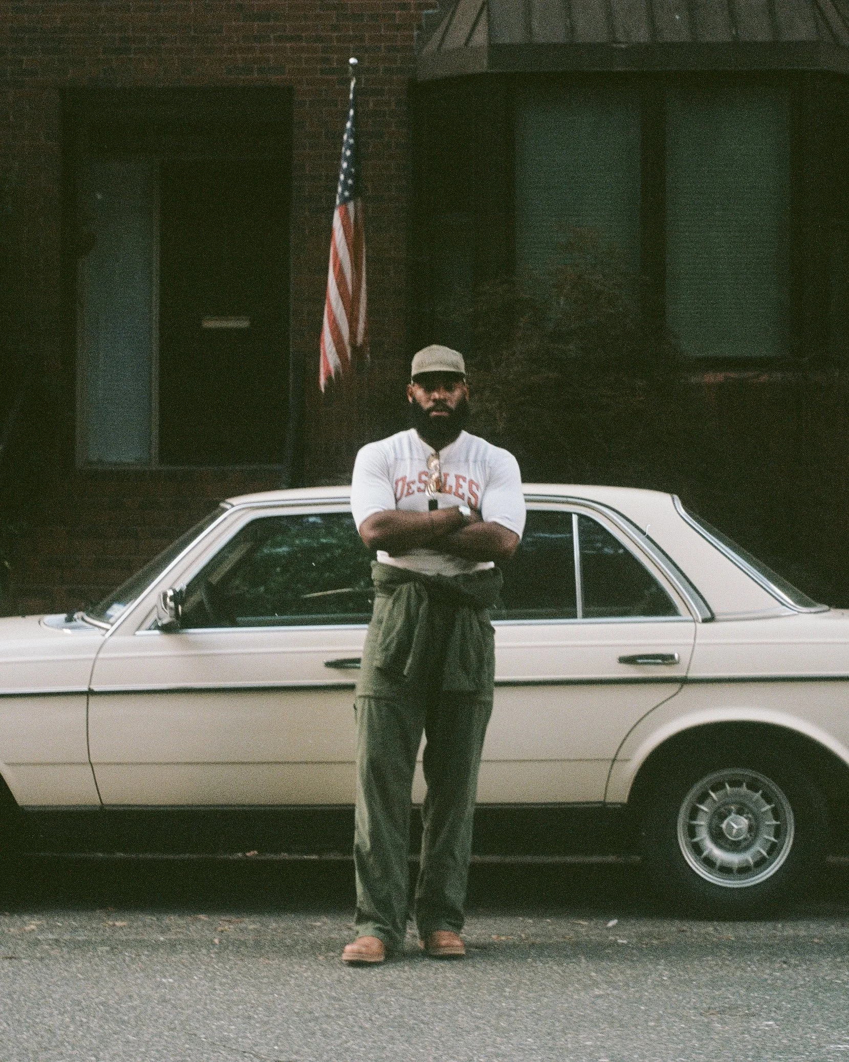 A man with a beard wearing a baseball cap, sunglasses, a white t-shirt with red text, green pants, and tan shoes, standing with his arms crossed in front of a vintage white car parked on a city street. An American flag is visible in the background, a