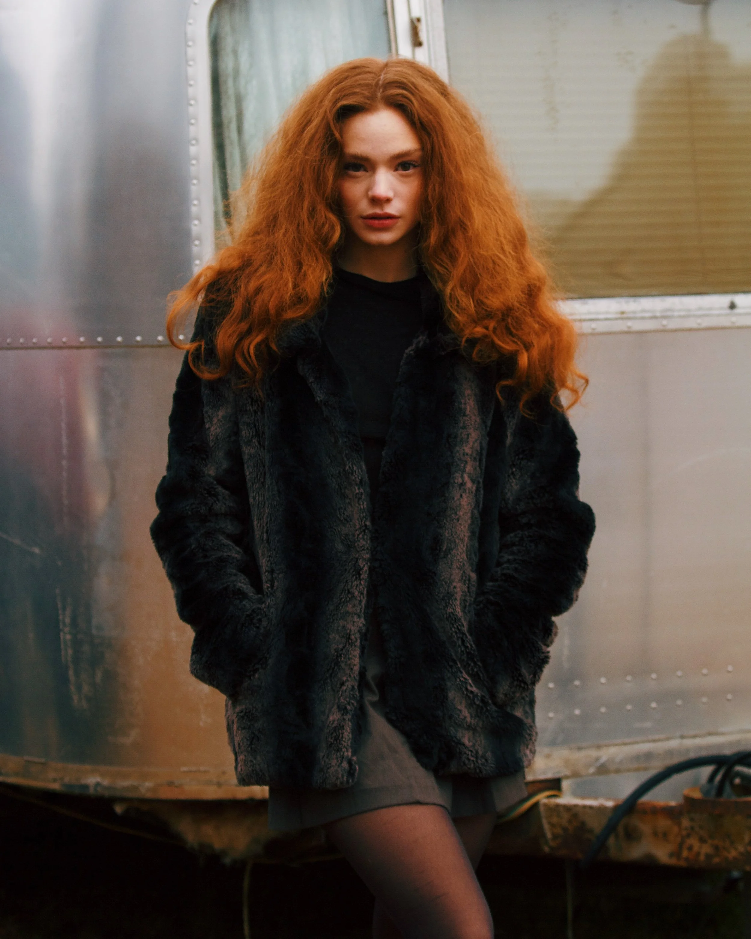 A young woman with long, curly red hair standing outdoors, wearing a black fur coat, black top, and gray skirt, with her hands in her coat pockets, in front of a metallic structure.