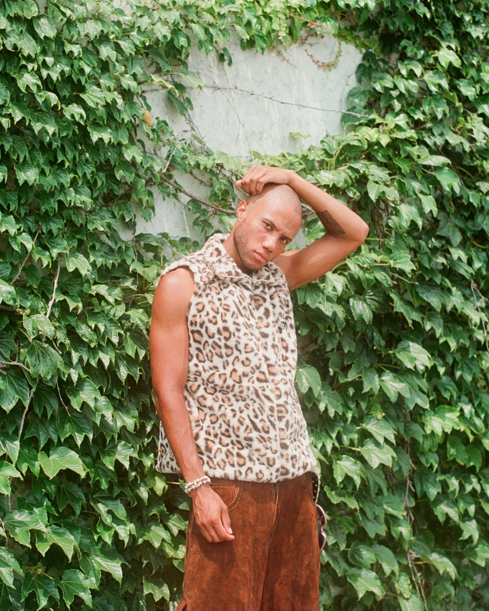 A man with a shaved head and tattoos, wearing a leopard print sleeveless vest and brown pants, standing in front of a wall covered with green ivy plants.