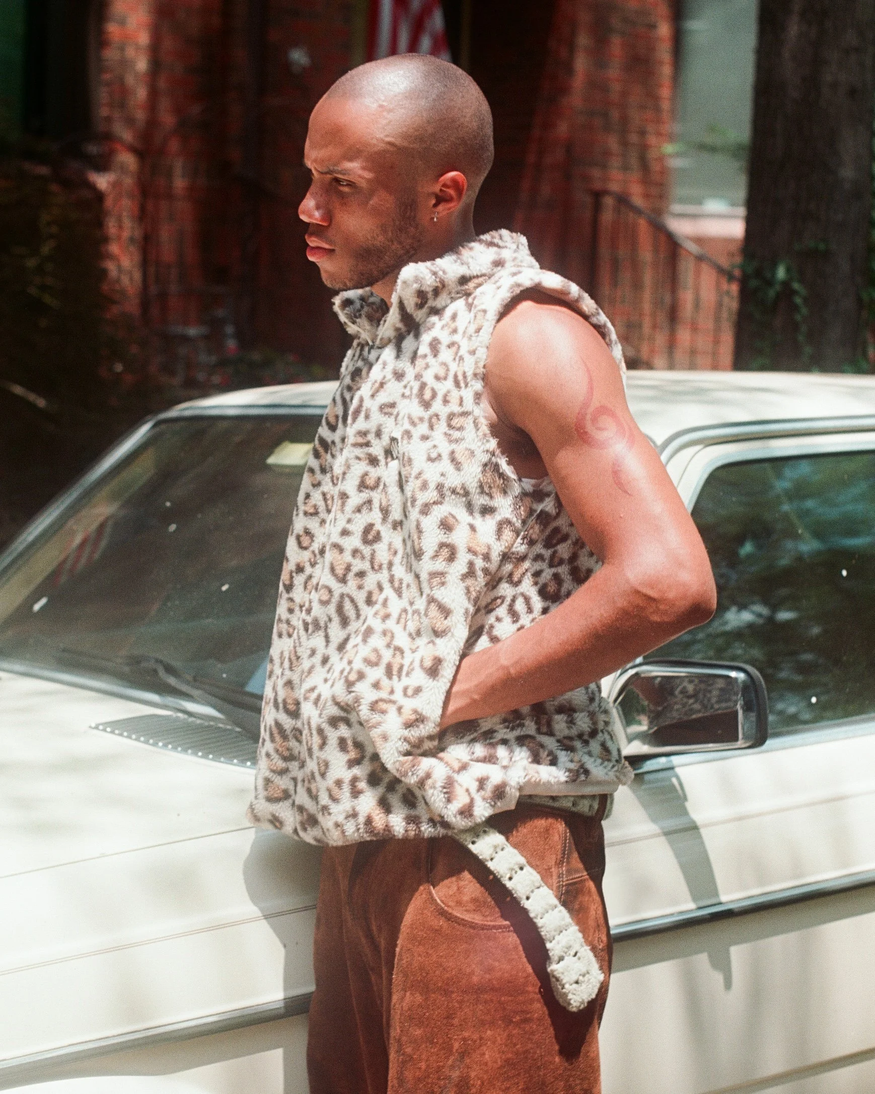 A person standing next to a white car, wearing a sleeveless leopard print shirt, brown suede pants, and a tail, with a tattoo on their upper arm, bald head, and a serious expression.
