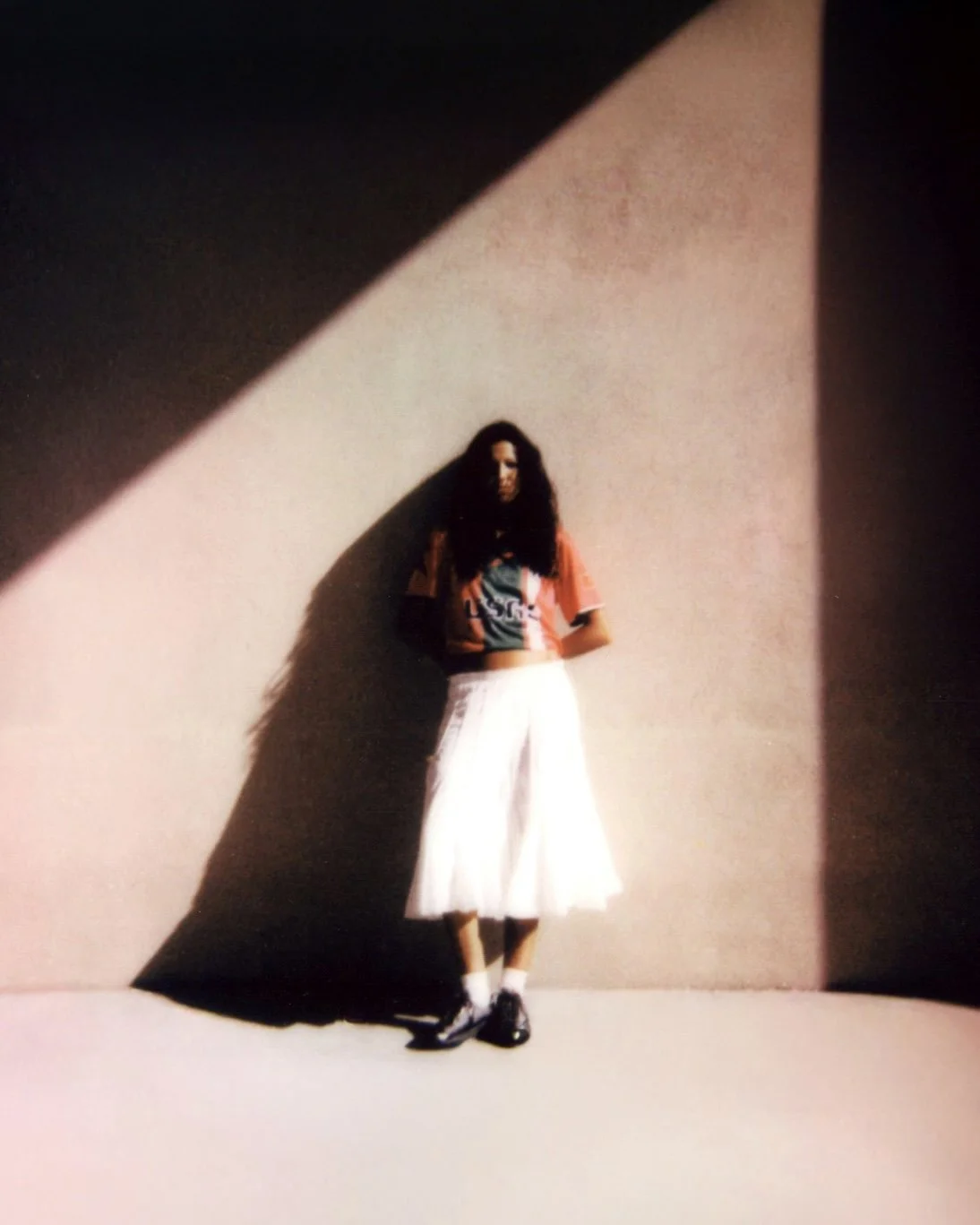 A woman standing against a wall with a large shadow cast behind her. She is wearing a colorful T-shirt, long white skirt, and black shoes, with her hands behind her back.