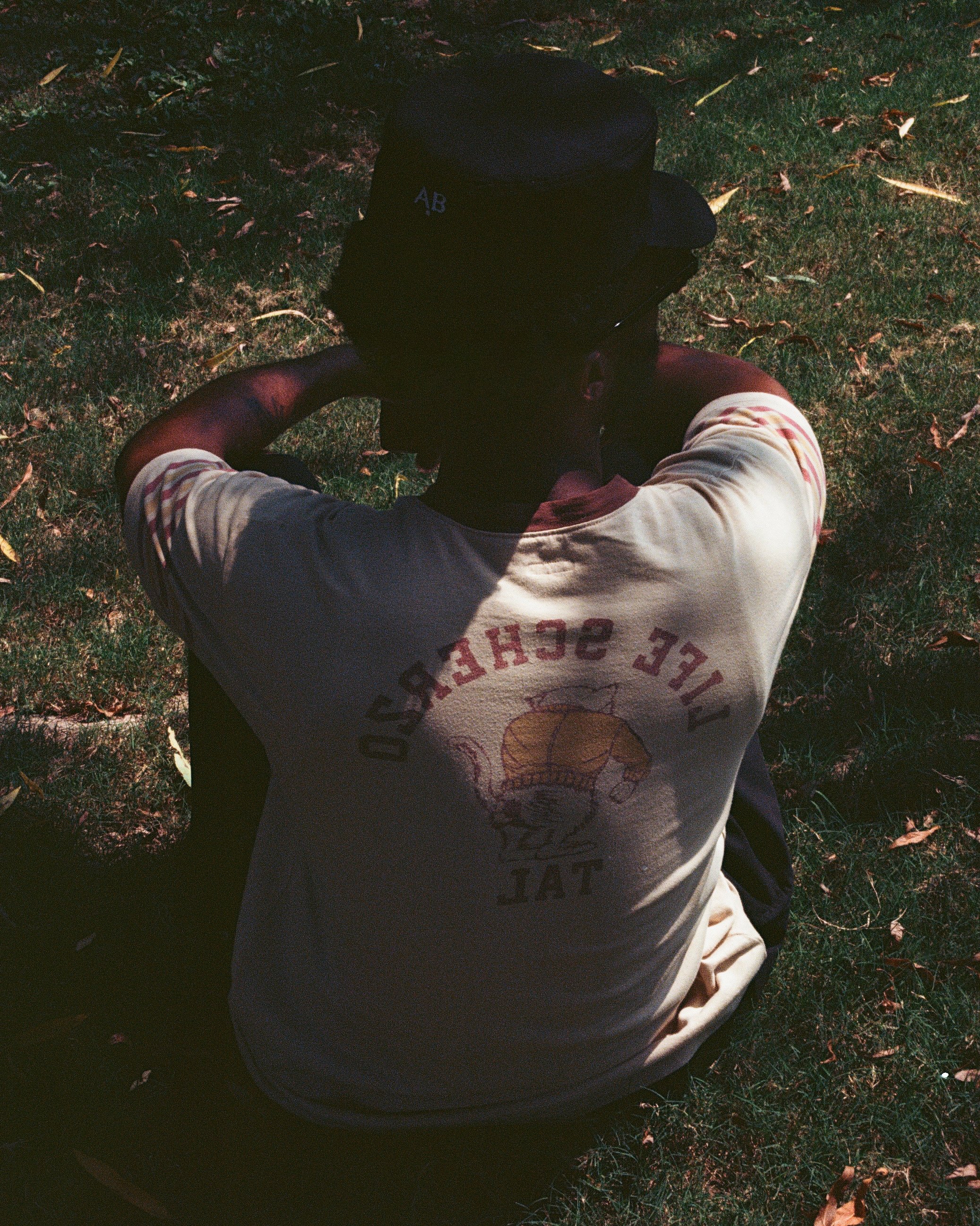 A person sitting outdoors on grass, wearing a beige T-shirt with a graphic and a black baseball cap with the initials 'AB' on it.