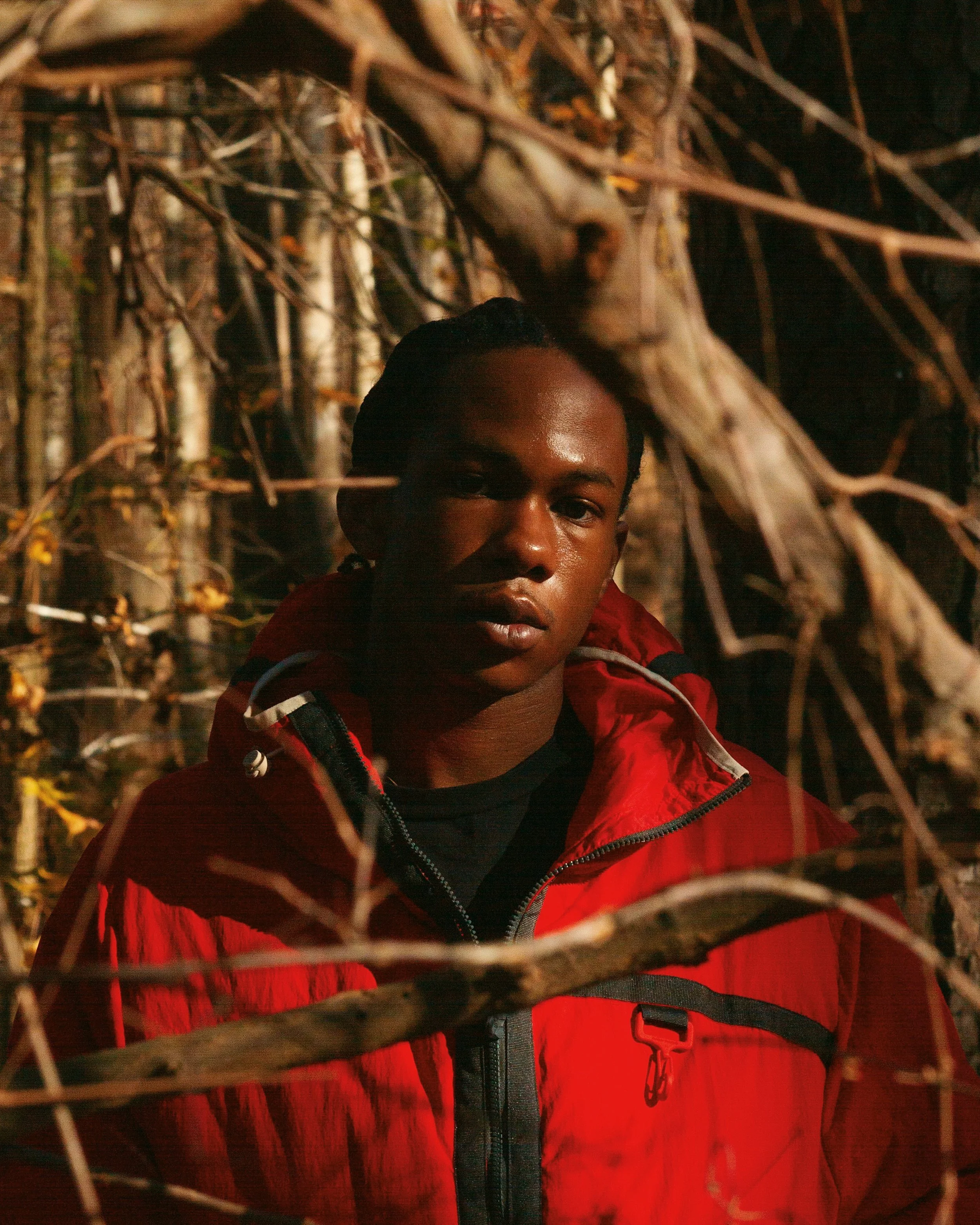 A person wearing a red jacket standing among tree branches in a forest, facing camera with sunlight highlighting face.