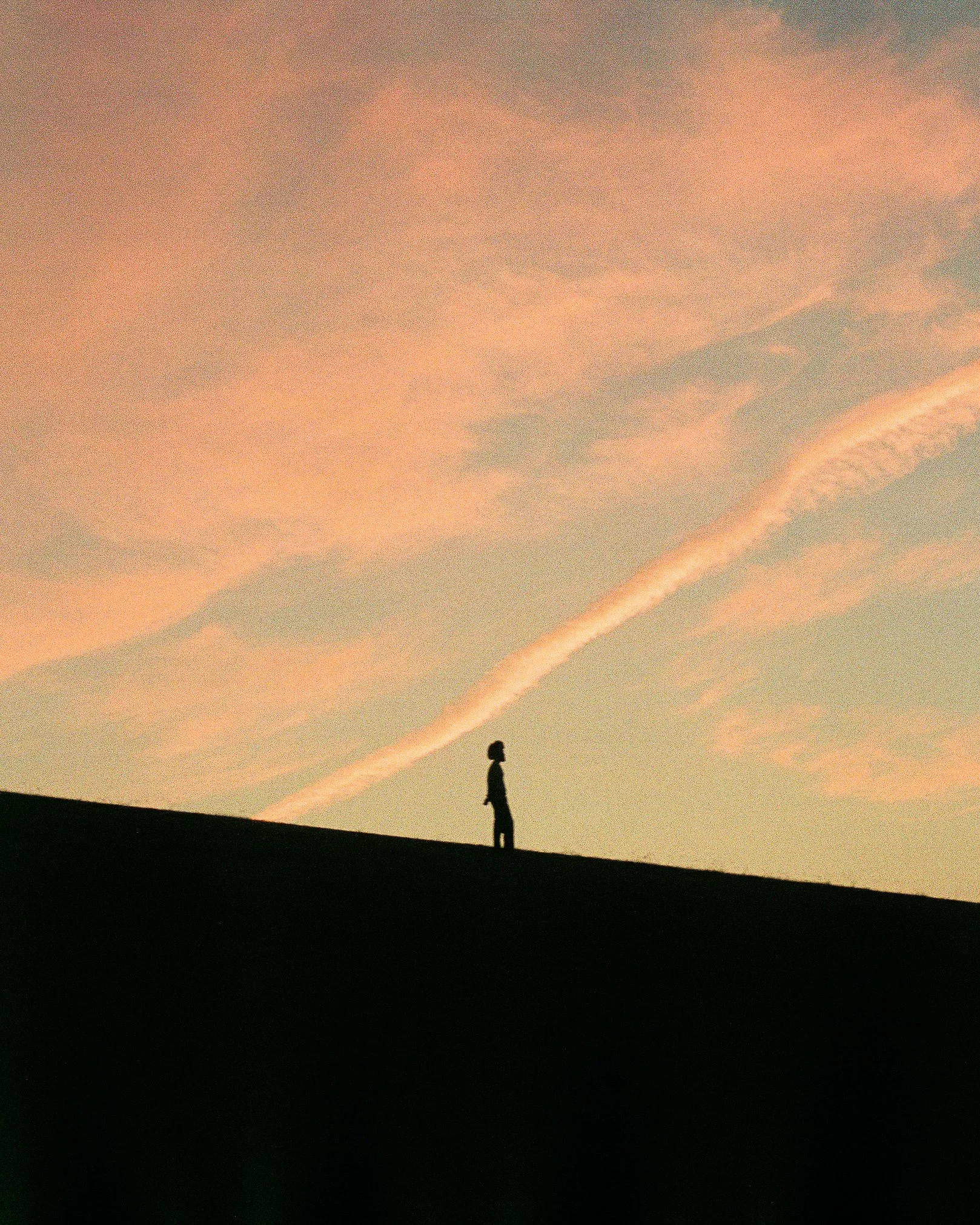 Silhouette of a person standing on a hill against a colorful sky with pink clouds and a streak of white clouds.