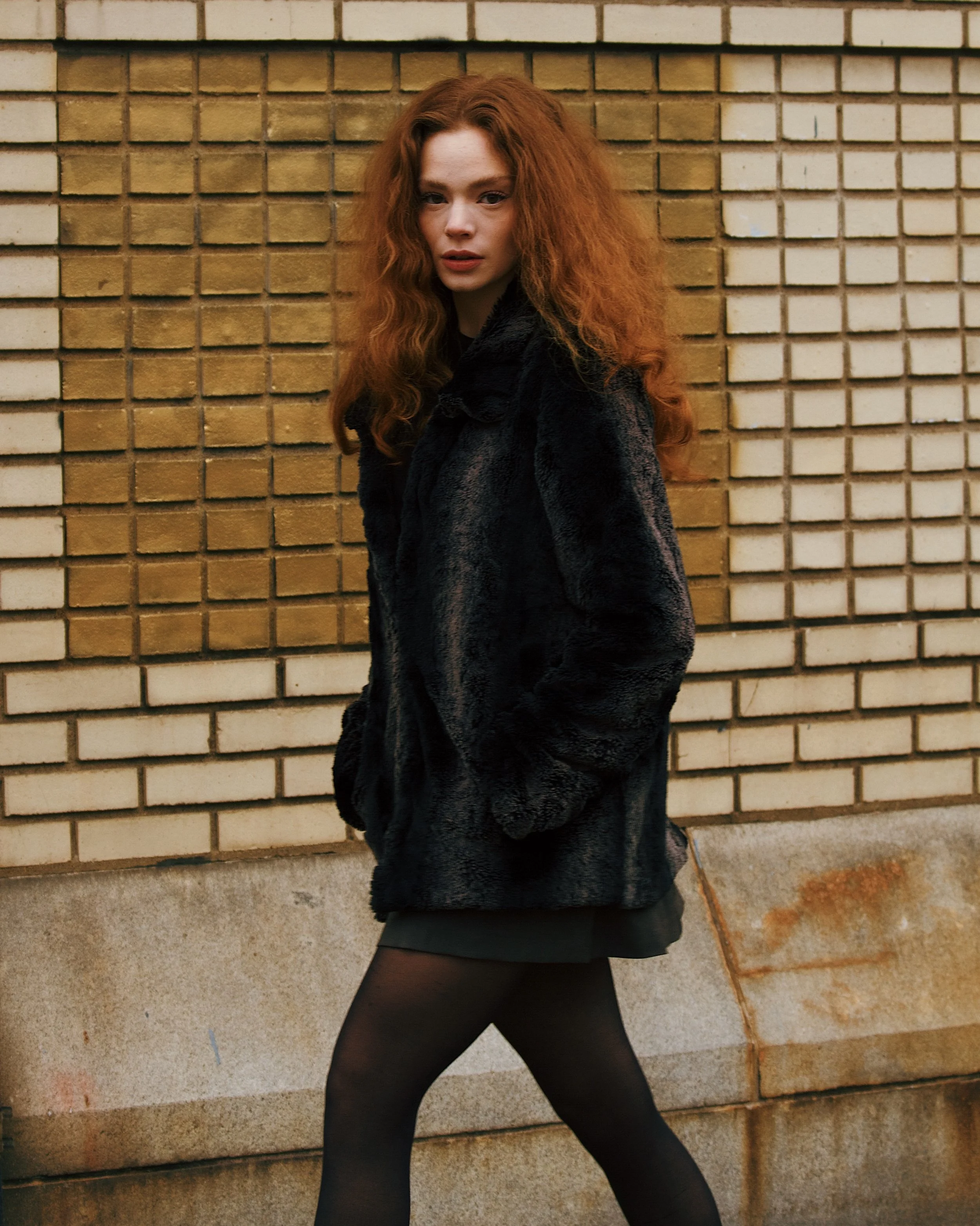 A young woman with long, curly red hair walking in front of a brick wall, wearing a black coat, black skirt, and black tights.