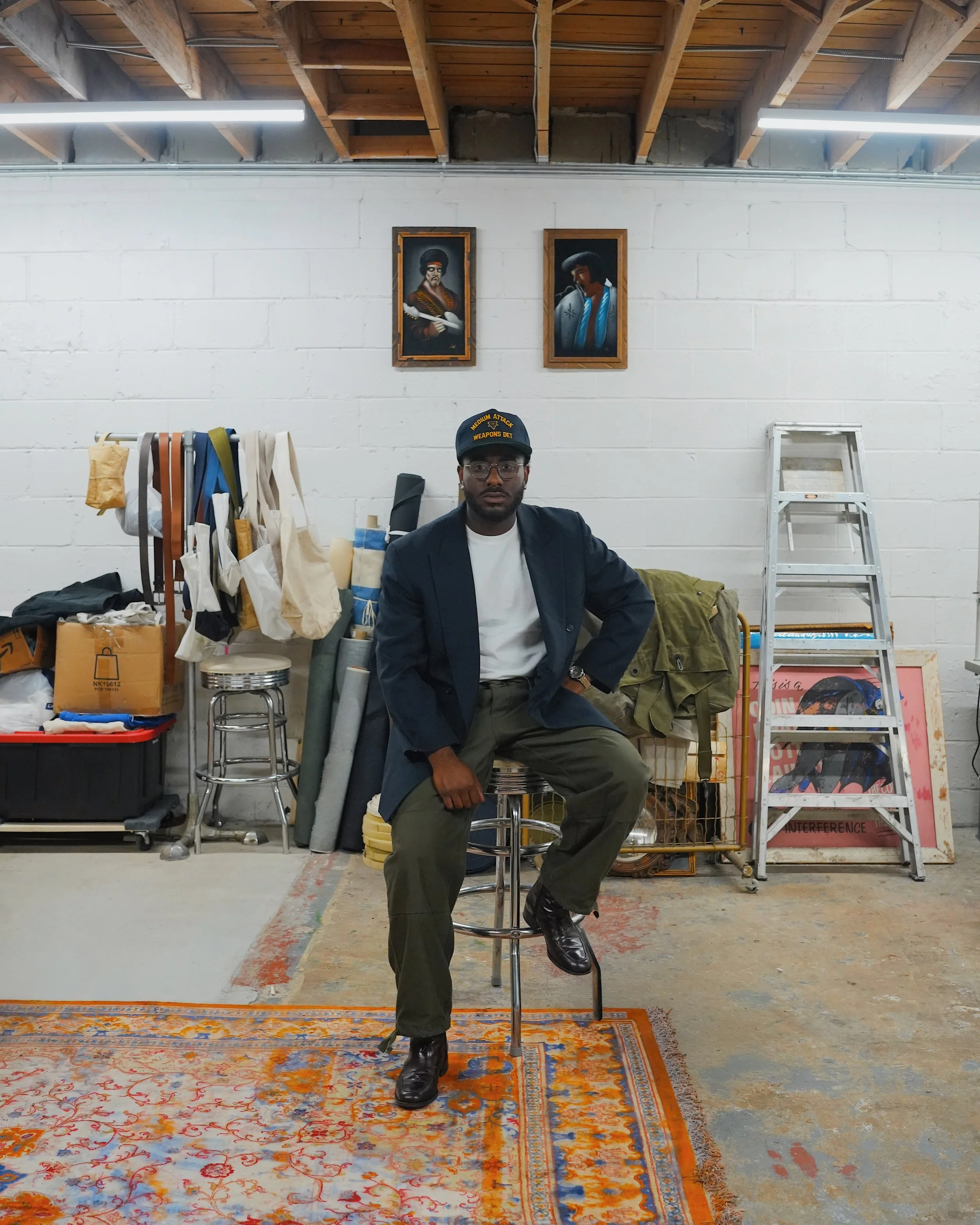 Man sitting on a stool in a storage room with paintings on the wall, ladder, boxes, and various stored items.