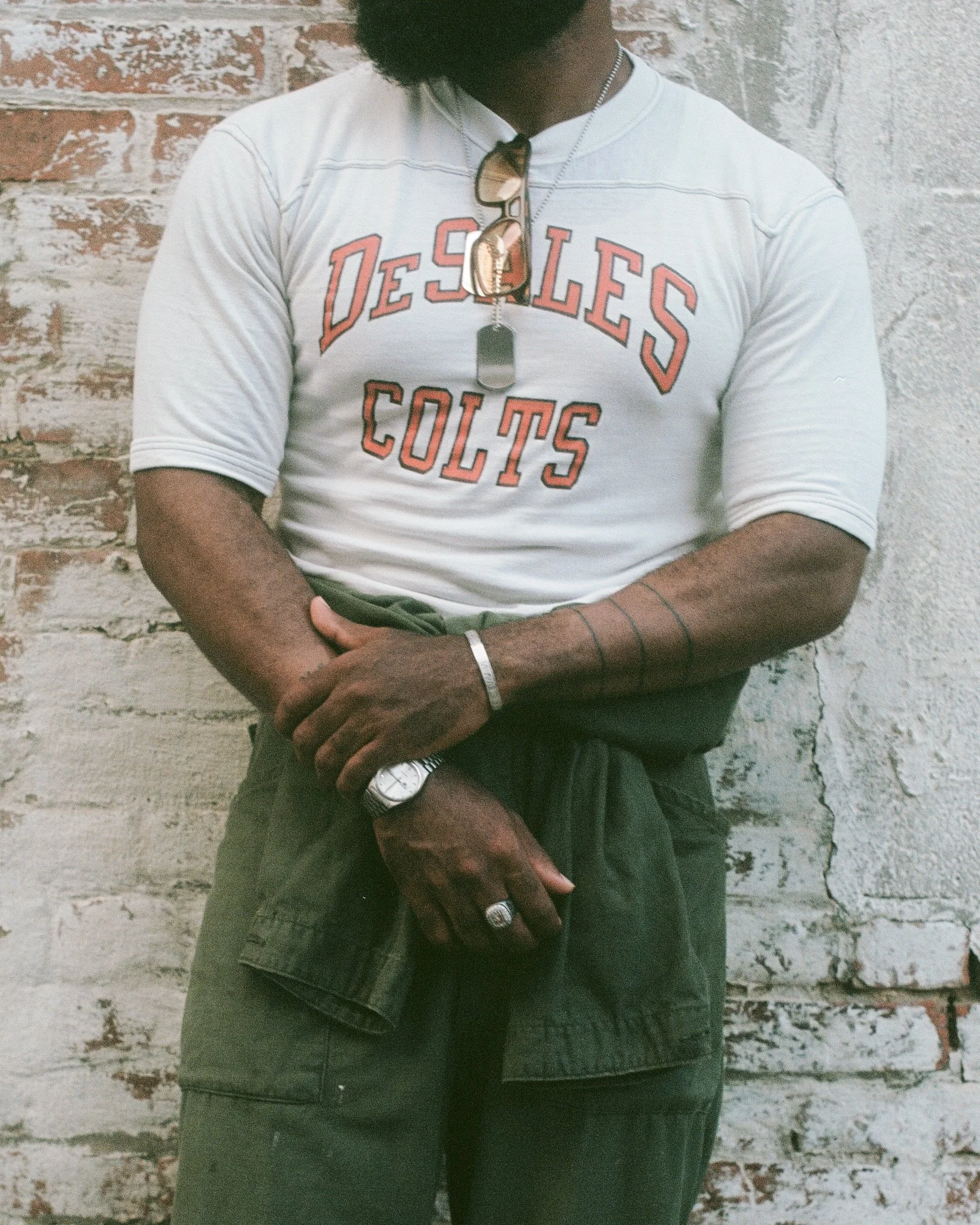 A man wearing a white t-shirt with 'Desoles Colts' printed on it, sunglasses hanging from his neck, and various rings and a watch. He is standing in front of a brick and textured concrete wall.