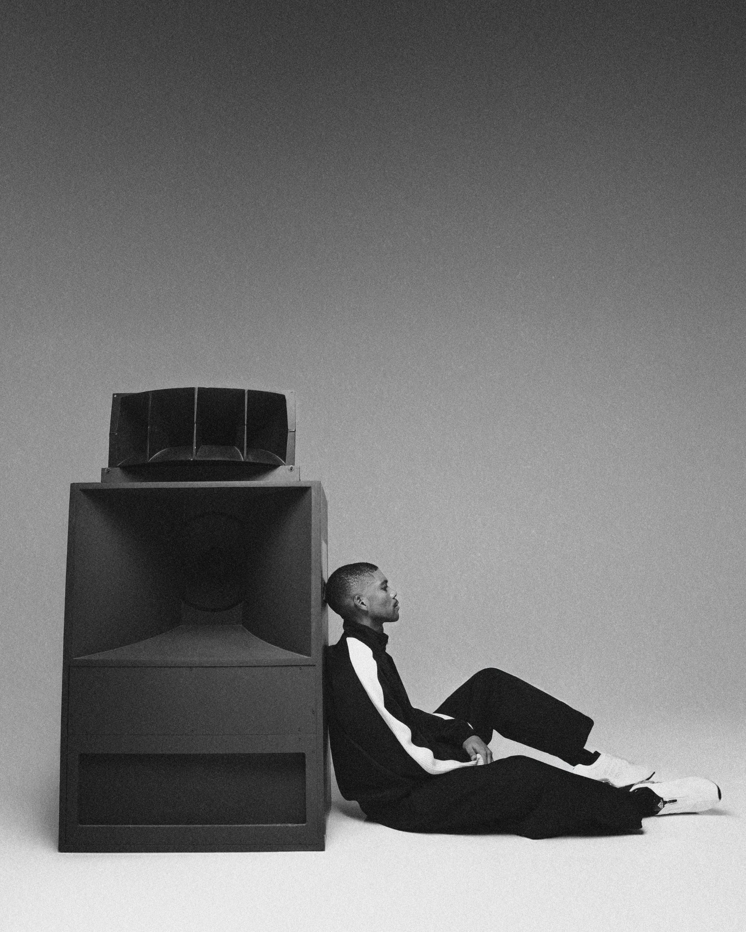 A person with a short haircut, dressed in a black and white tracksuit, sitting on the floor with legs extended, leaning against a large speaker, in a minimalist black and white studio setting.