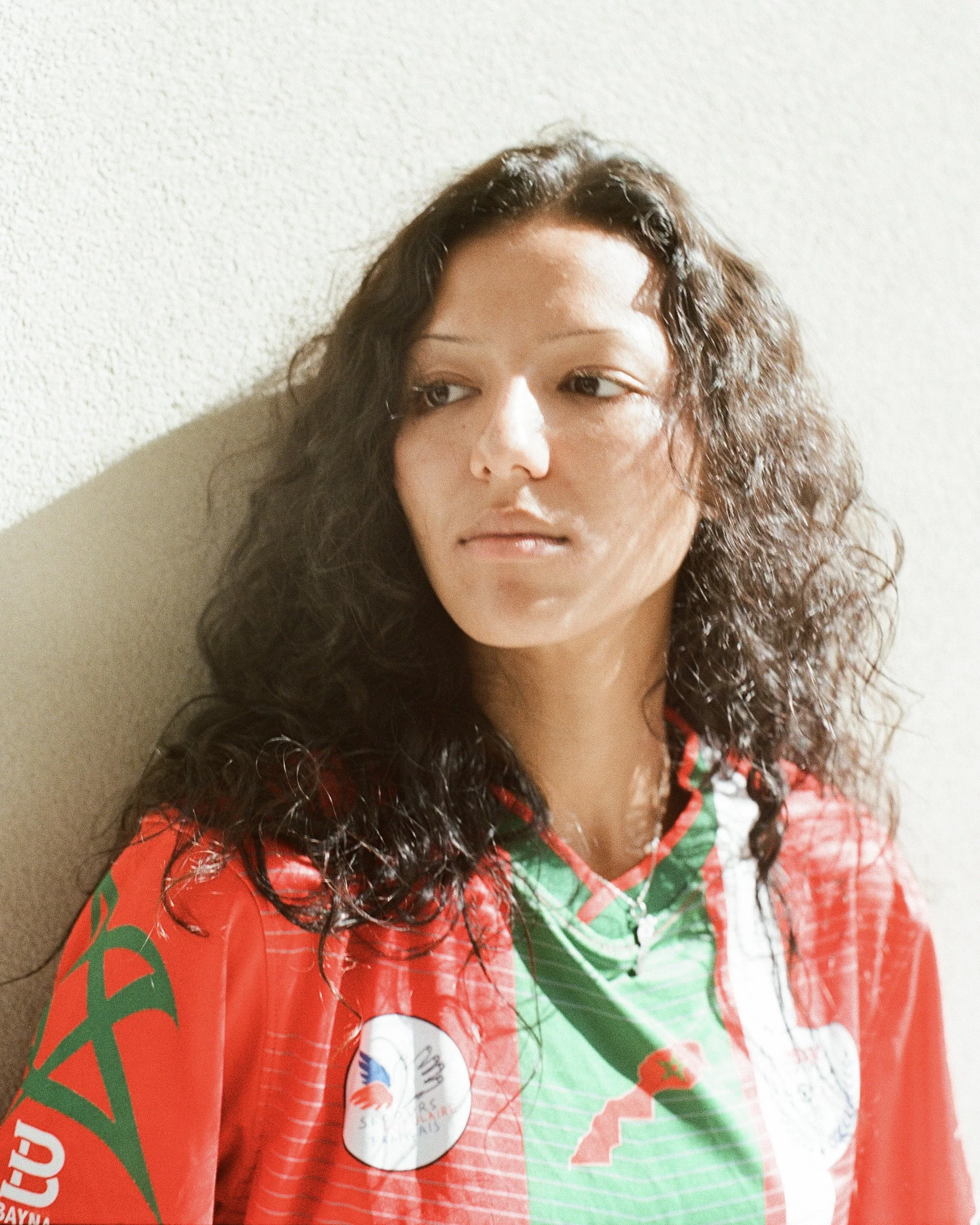 A woman with curly brown hair leaning against a textured beige wall, wearing a red and green sports jersey with patches and logos.