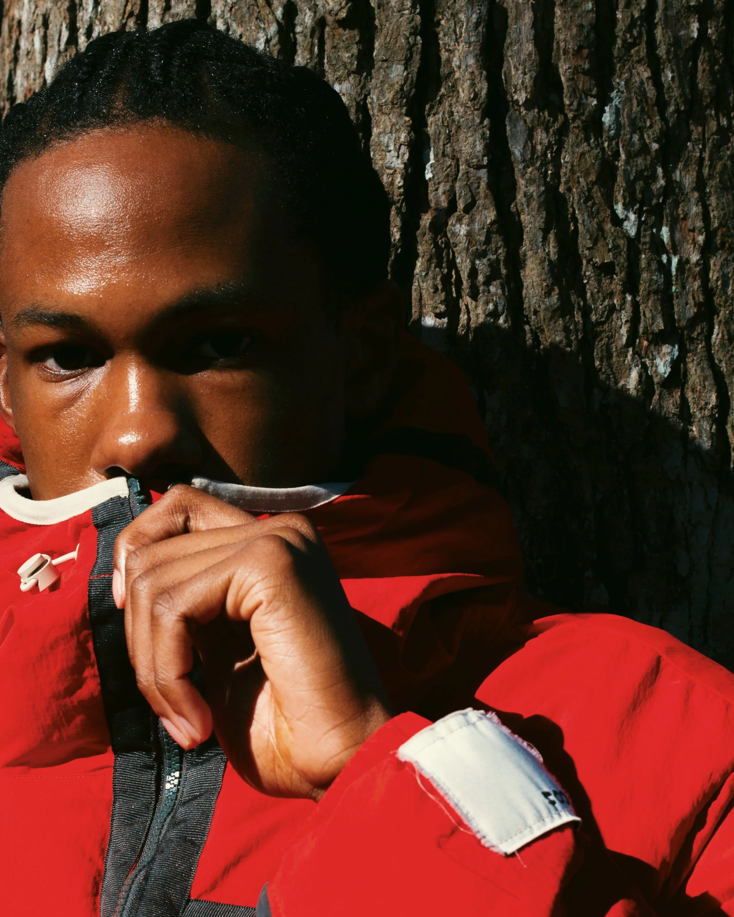 A person in a red jacket standing in front of a tree trunk, holding the collar of their jacket near their face, with the sunlight highlighting their face and the rough texture of the tree bark in the background.