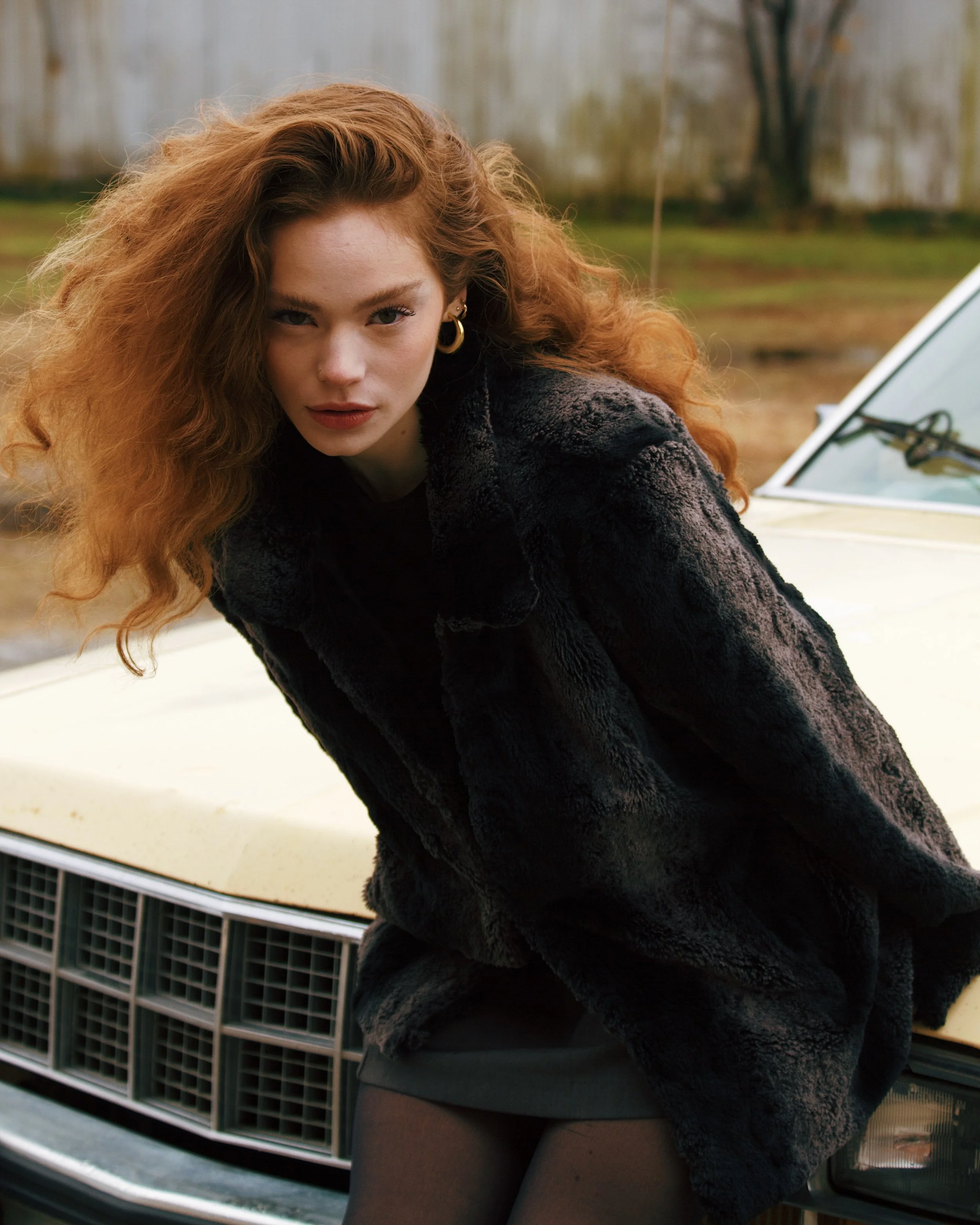A woman with long red curly hair leaning on the hood of a beige vintage car, wearing a dark faux fur coat and black tights, outdoors with a blurred fence and trees in the background.